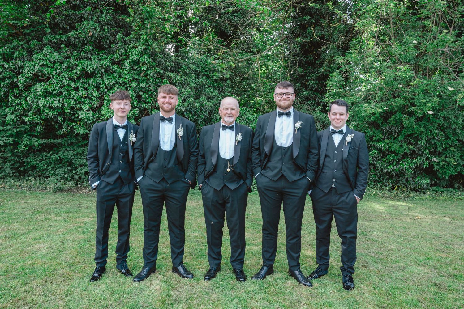 Five groomsmen in black tuxedos with bow ties pose outdoors on green grass with lush trees behind them.