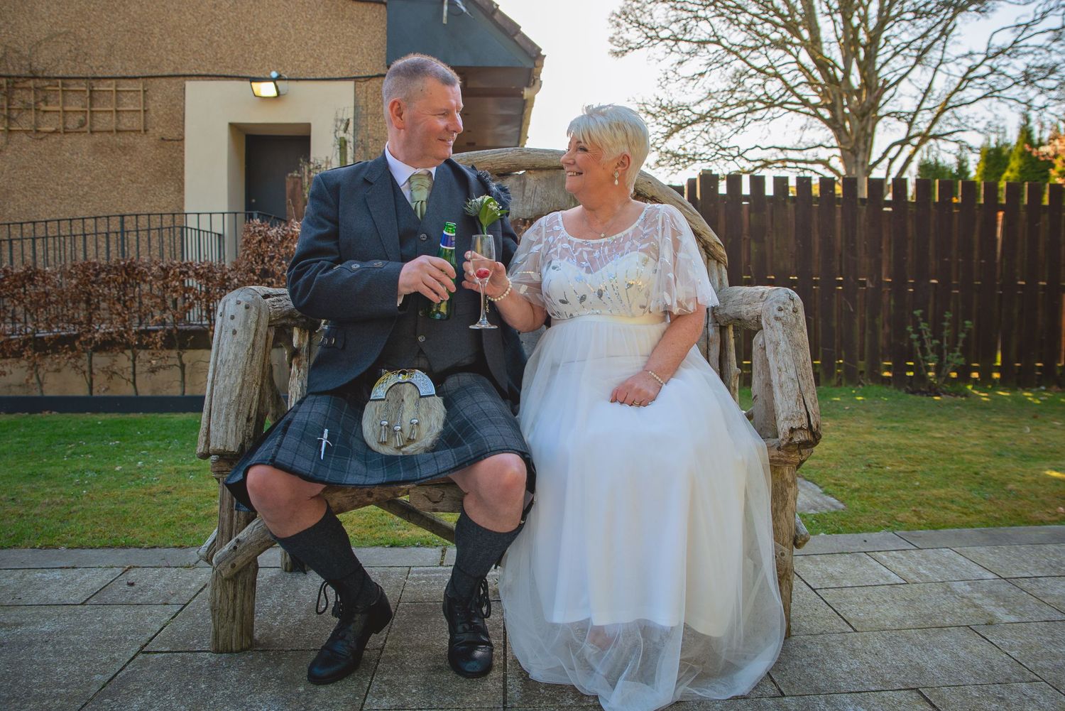 Couple in Scottish wedding attire sitting together on a wooden bench in a garden.