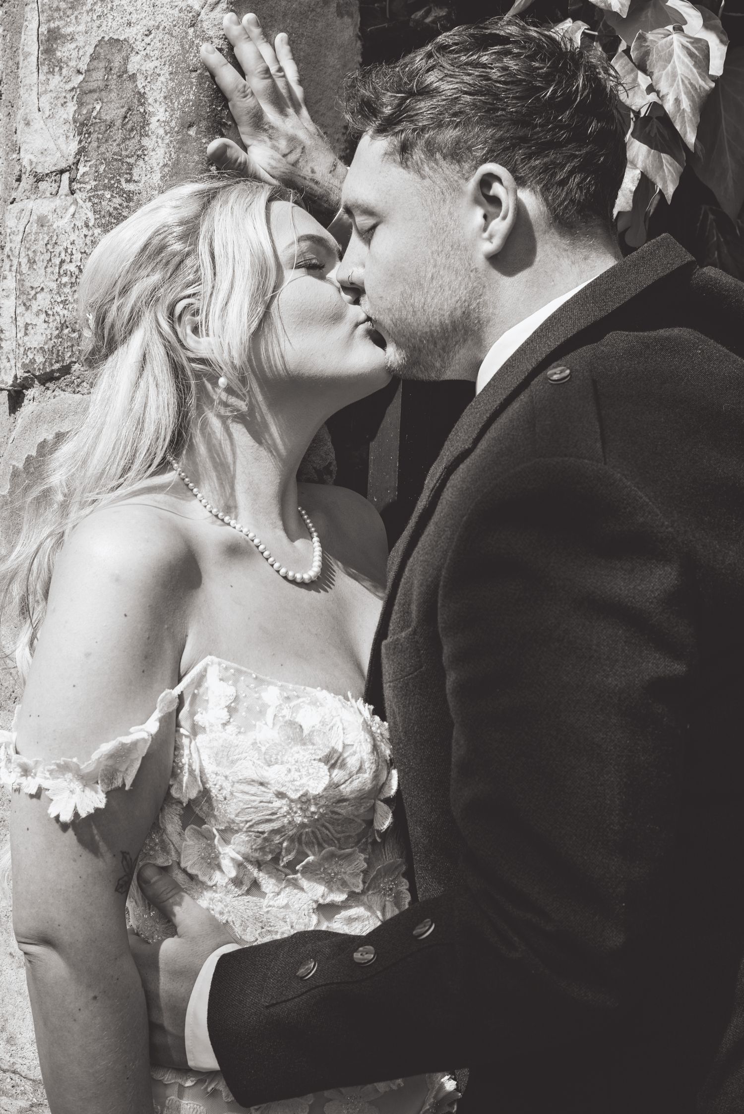 A romantic black and white wedding photo of a couple sharing an intimate kiss against a stone wall.