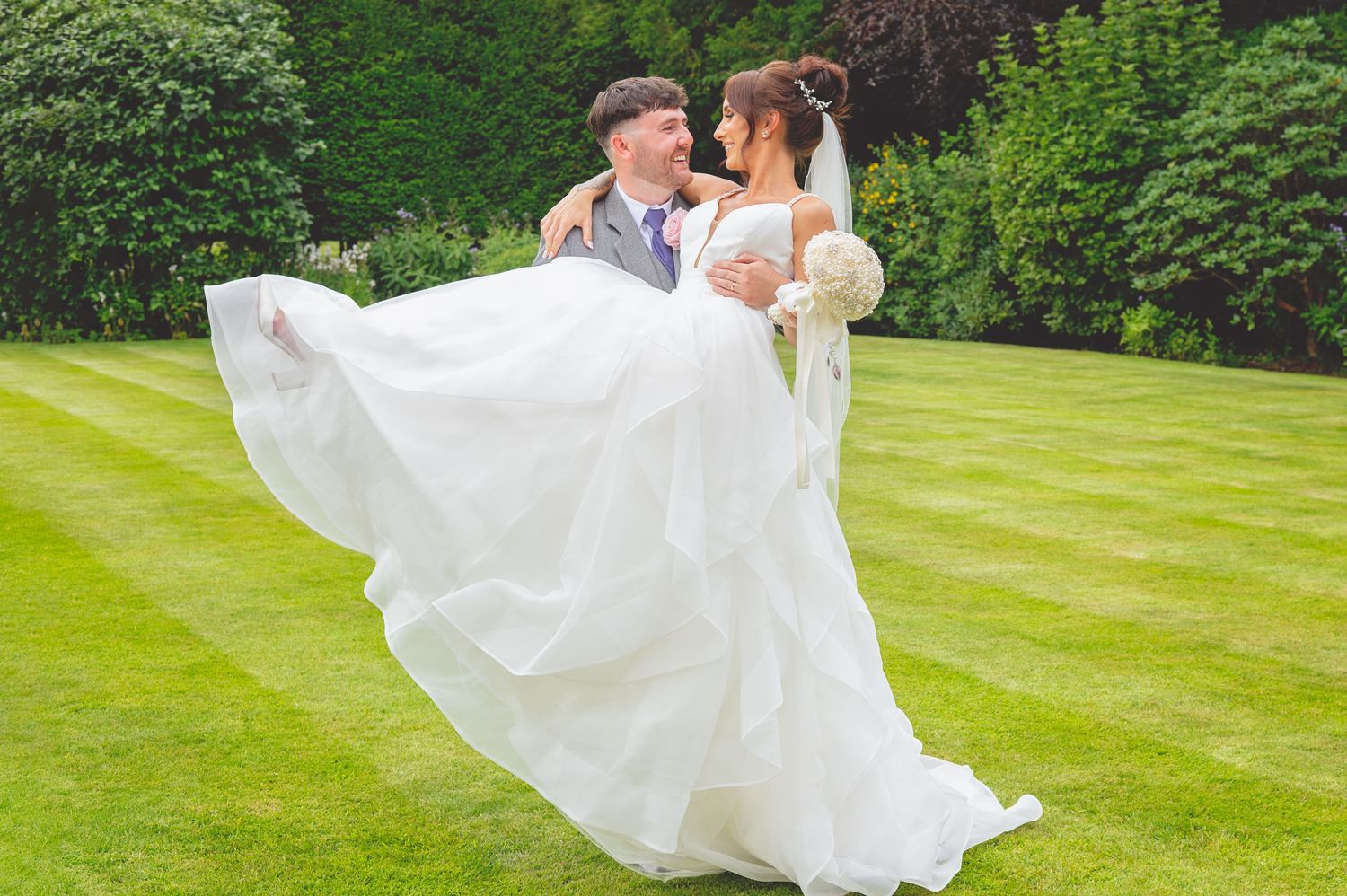 Wedding couple on green lawn with flowing white dress being lifted in romantic pose.
