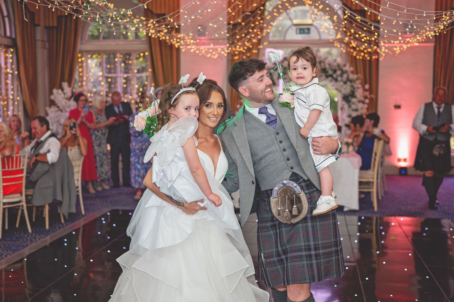 Wedding family portrait with couple and children under fairy lights at reception venue.