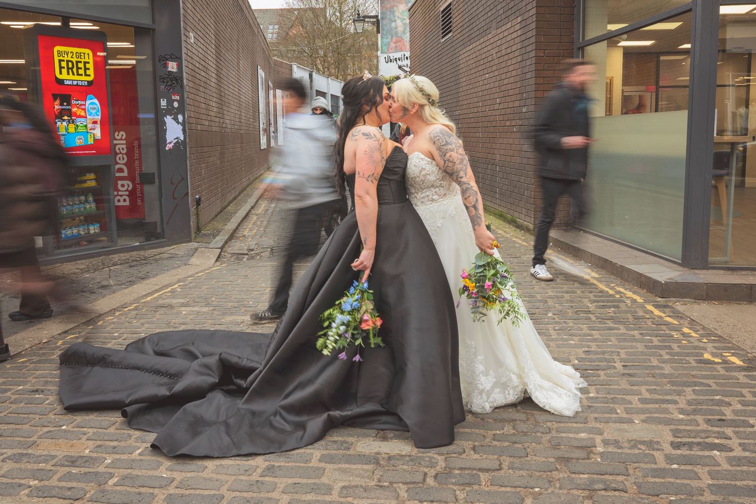 Two brides share a kiss on a cobblestone street, one in a black gown with train, the other in white, holding bouquets.