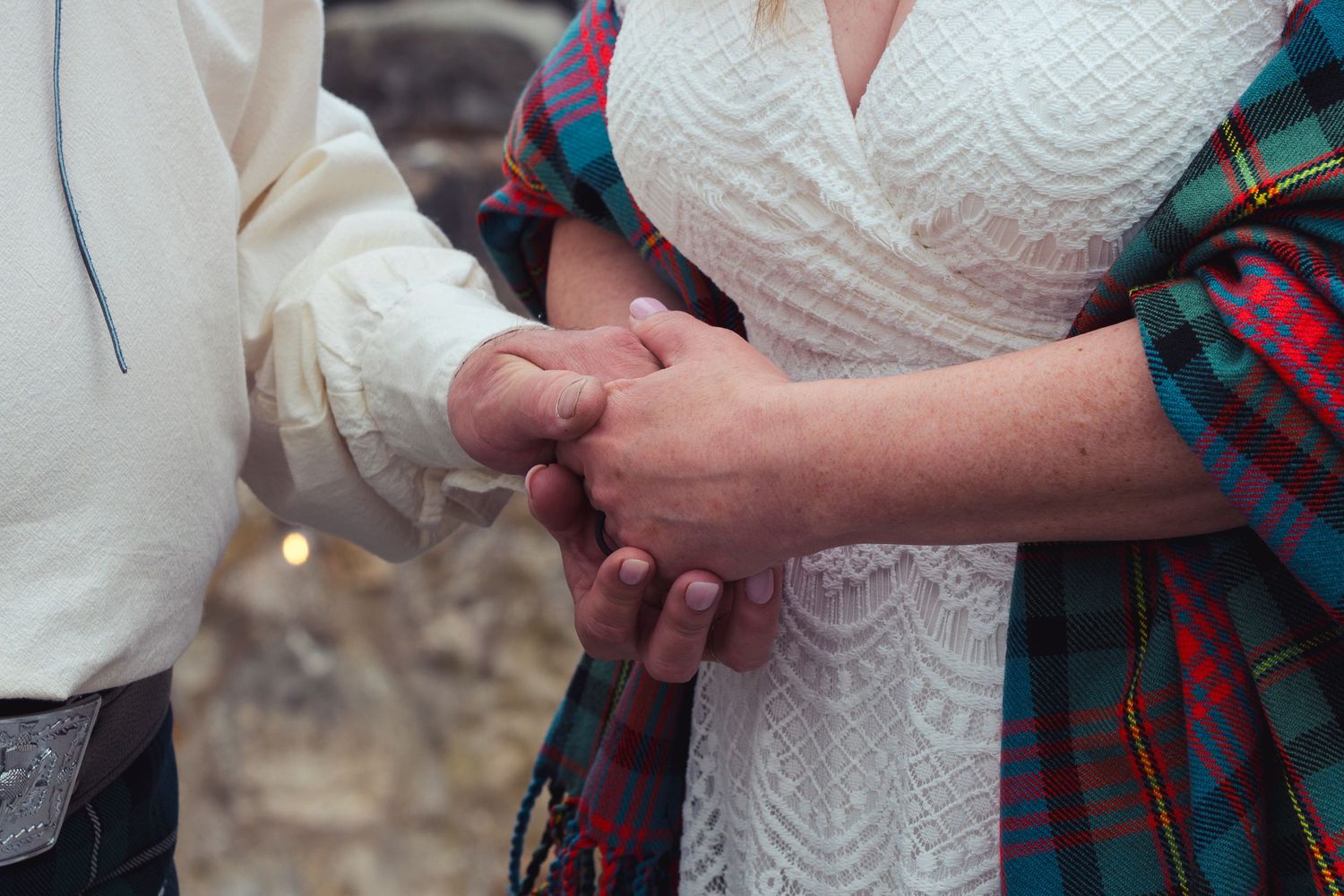 Couple holding hands at wedding, bride in lace dress with red tartan plaid sash in romantic outdoor ceremony.