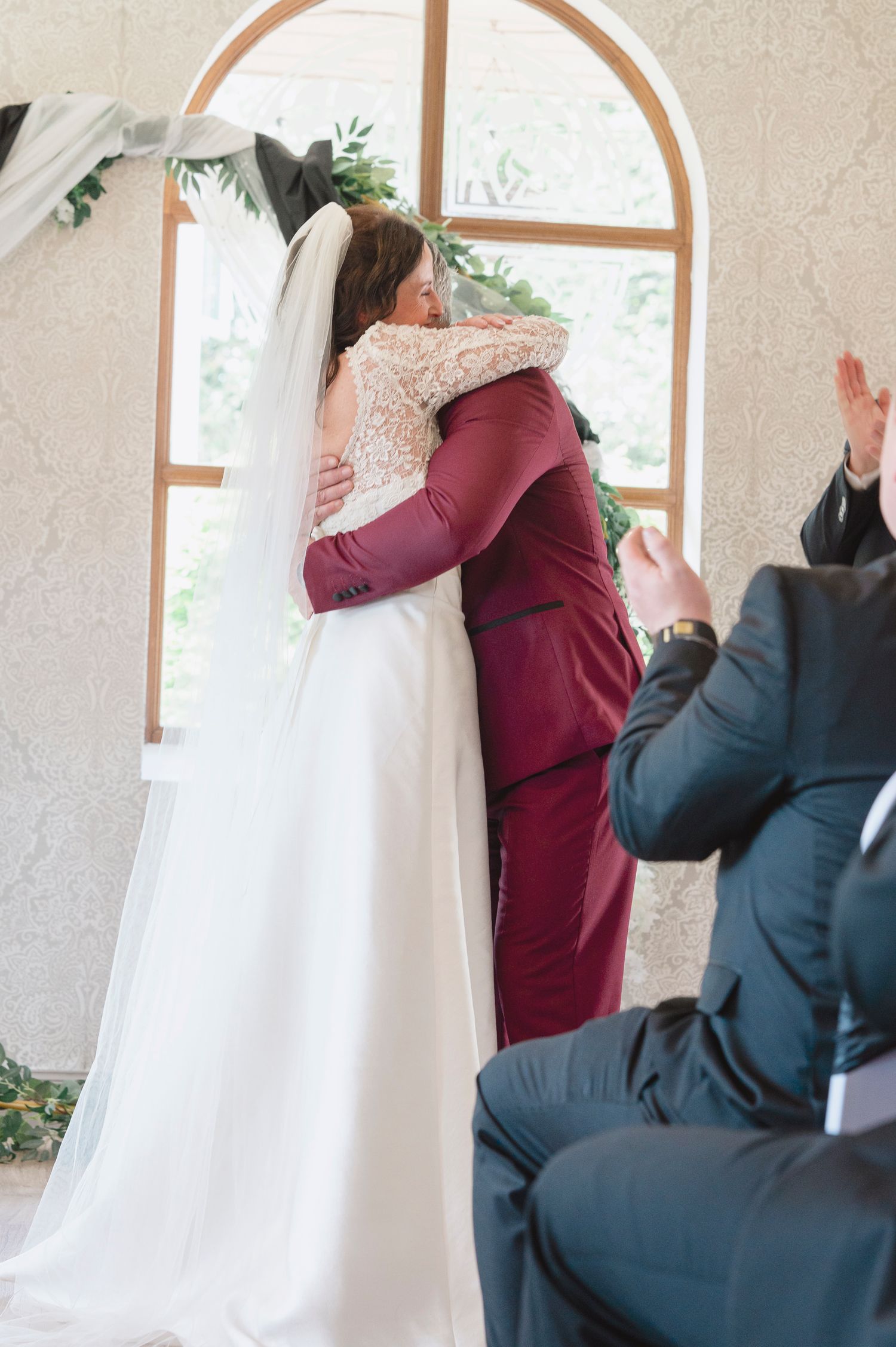 Bride and groom share a heartfelt hug at the wedding altar, with guests applauding in the foreground.