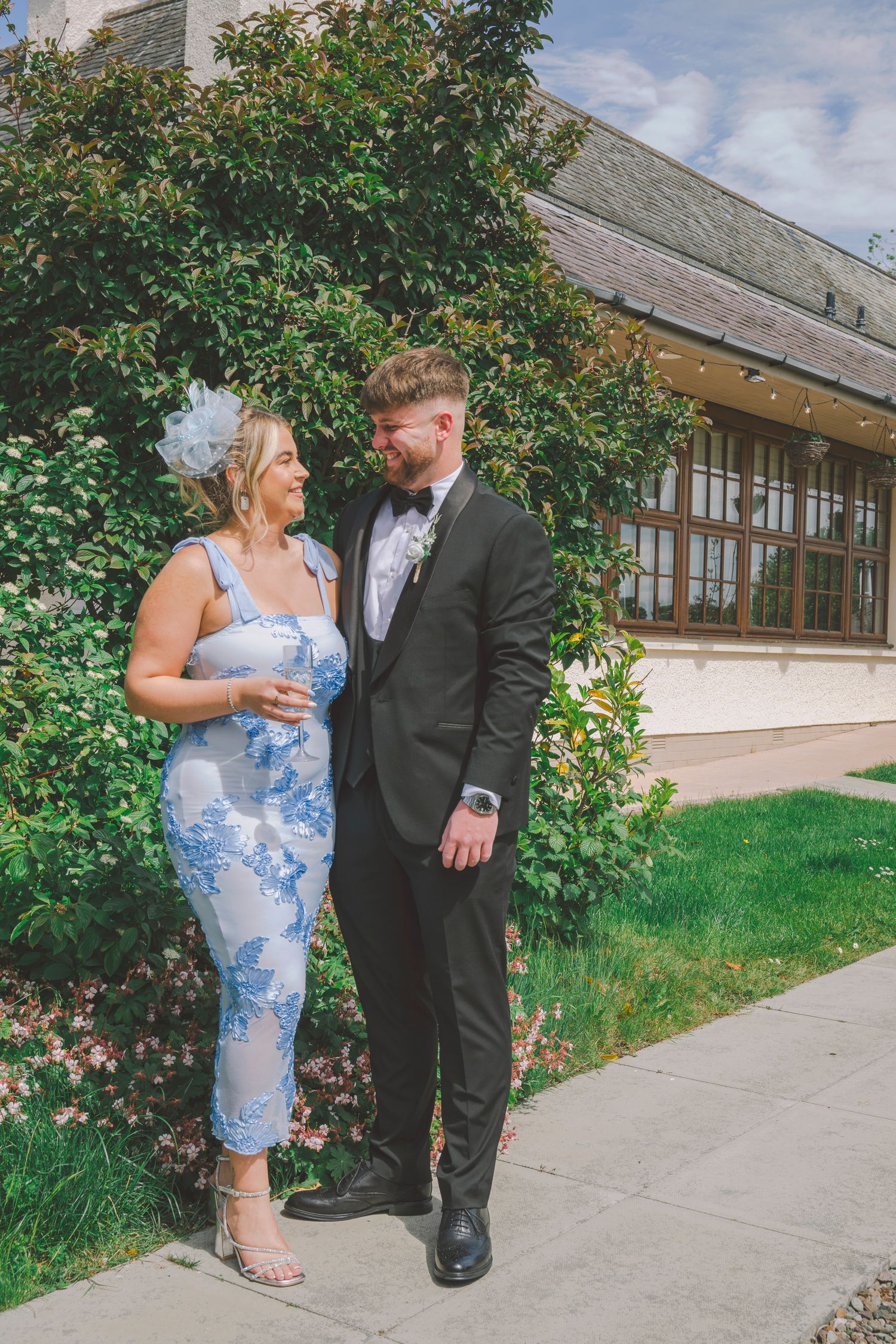 A couple dressed in formal attire stand together outdoors, smiling, with lush greenery and a building behind them.