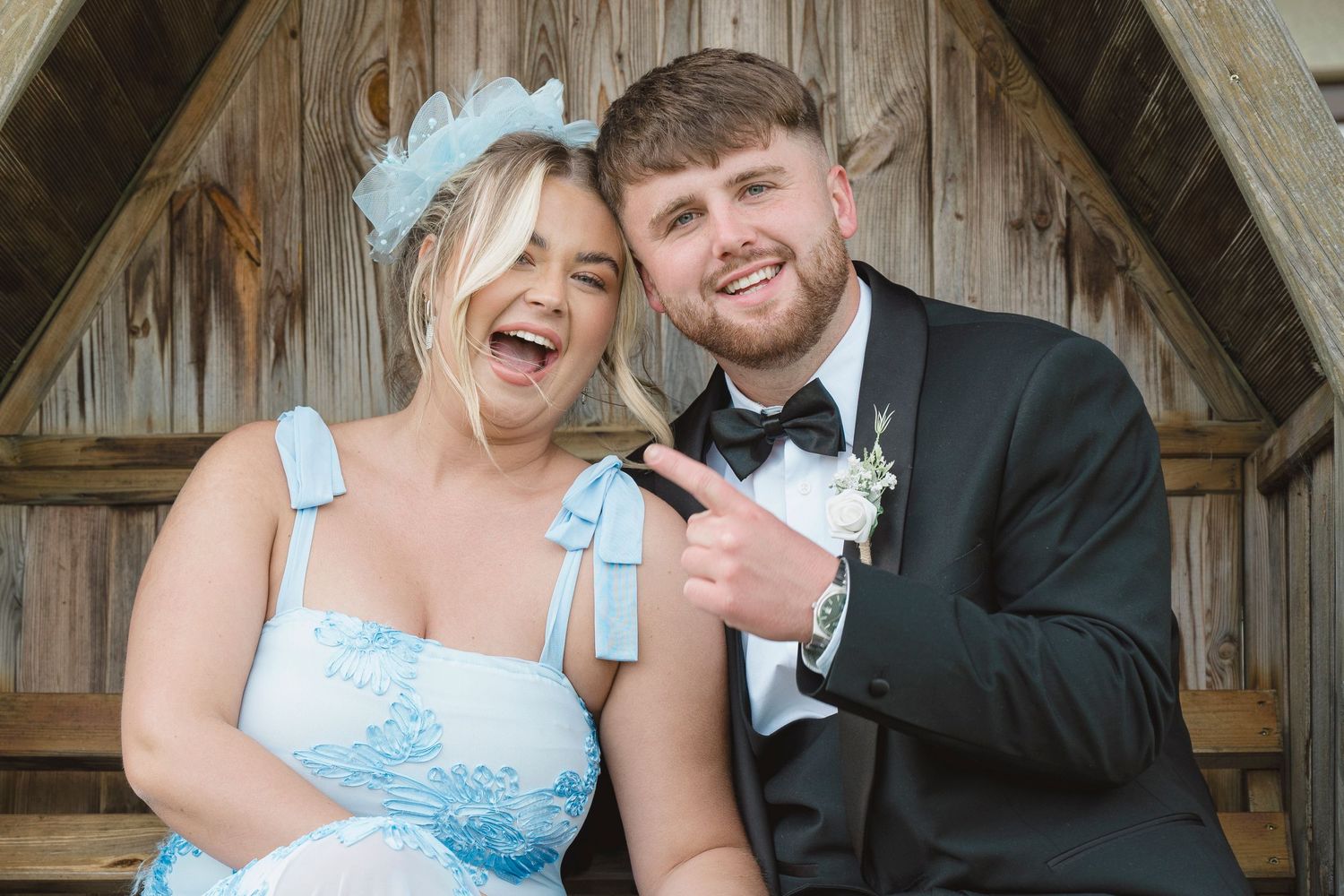 Couple in formal attire smiling at prom, woman in blue lace dress with bow, man in black tuxedo with boutonniere.