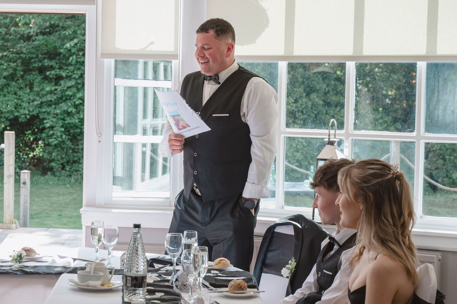 Man in vest and bow tie giving speech at elegant wedding reception with guests seated at table.