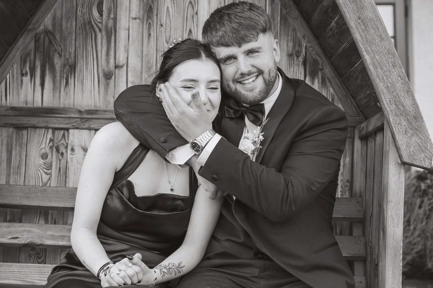 Black and white photo of a laughing couple in formal wear sitting together on wooden steps.