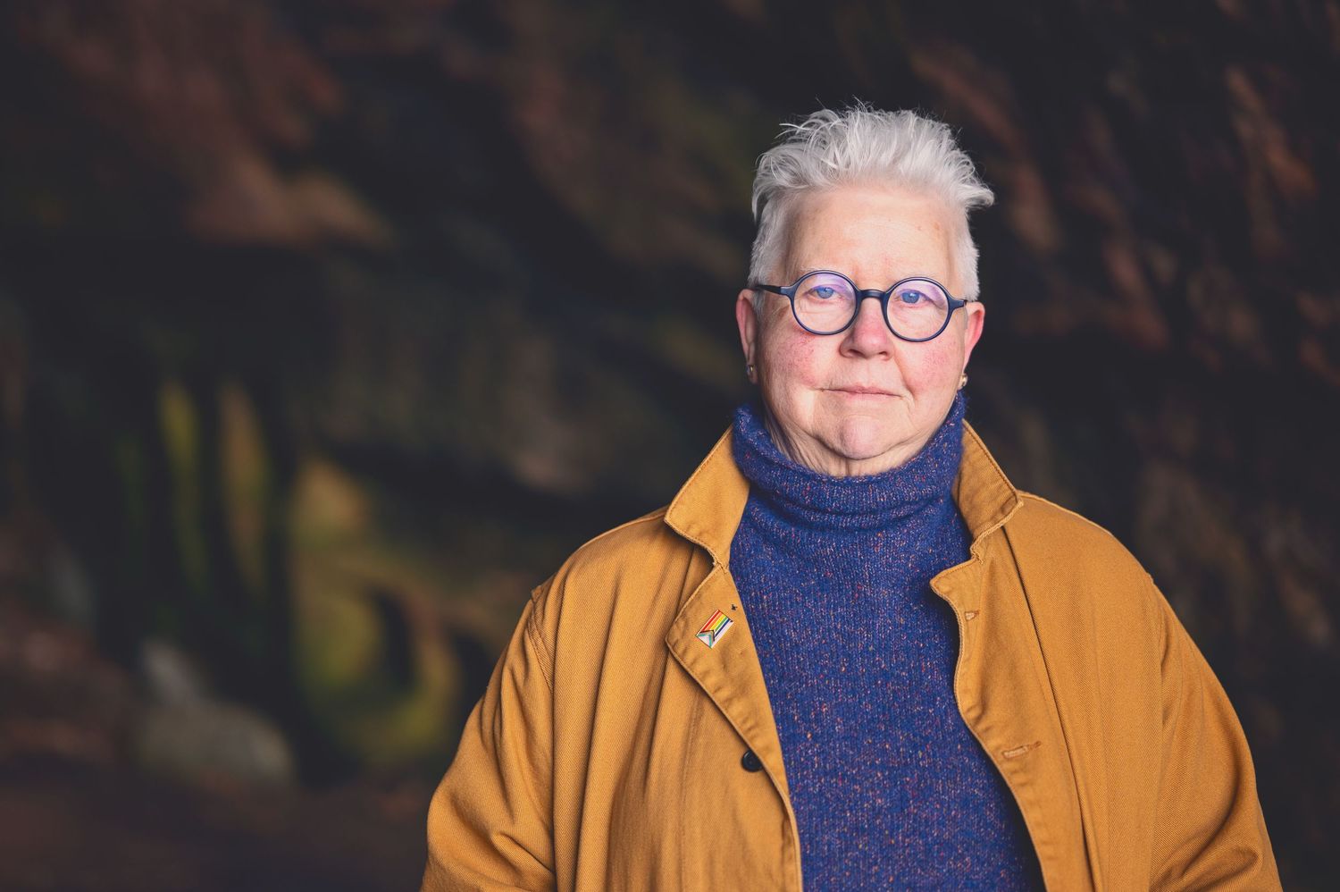 The Author Val Mcdermid with short white hair and round glasses wearing a mustard jacket and blue turtleneck sweater in the Weymss caves
