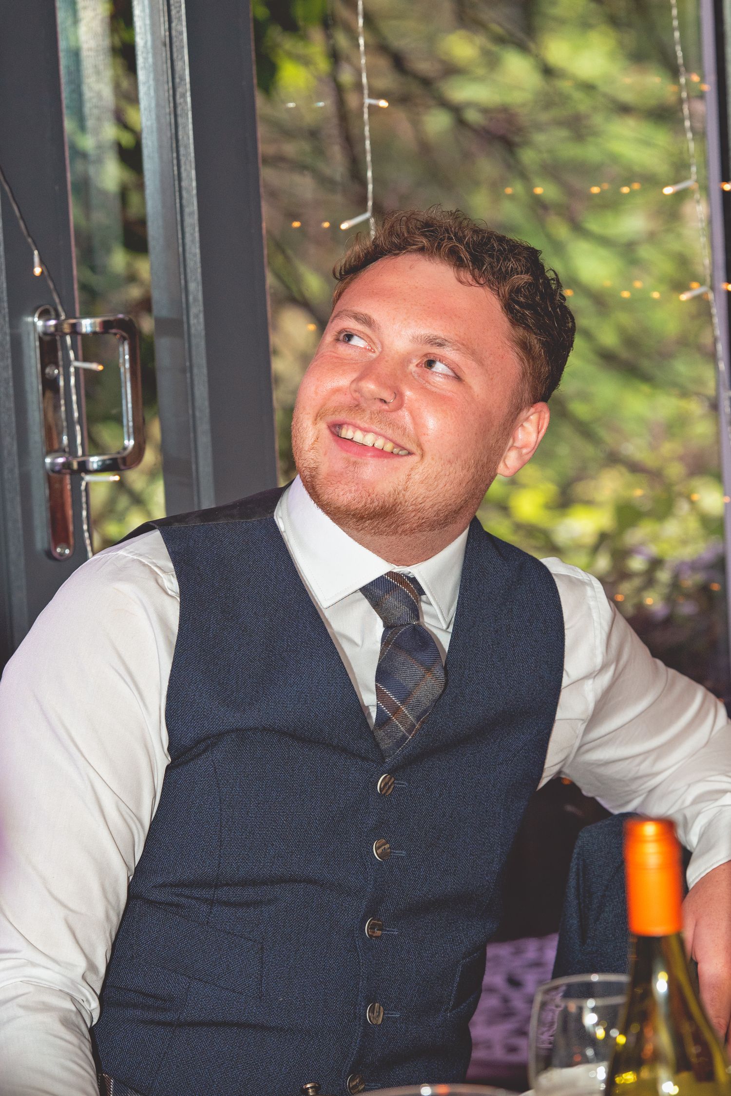 A guest in a navy waistcoat smiles while relaxing at a wedding reception.