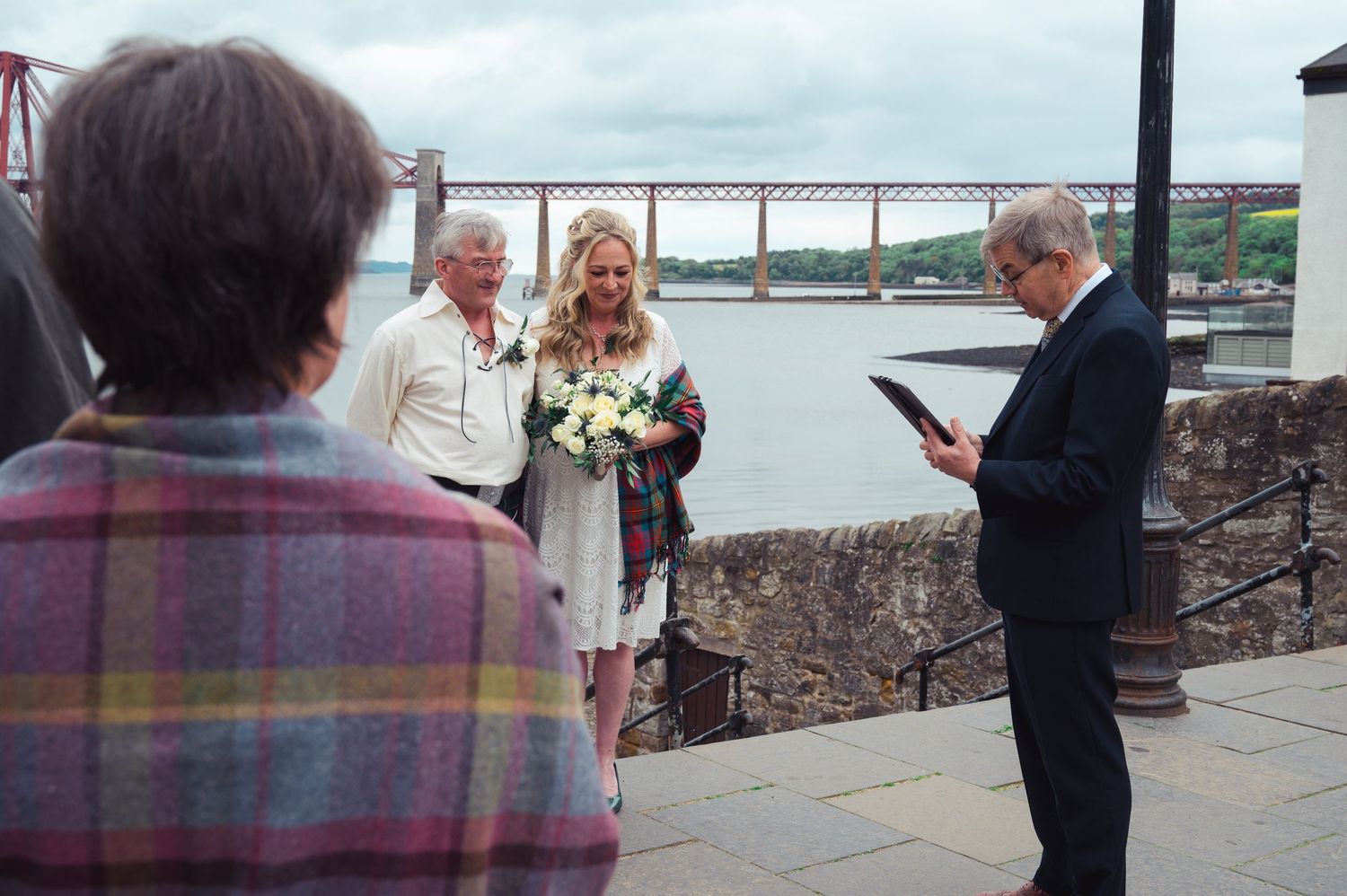 Outdoor wedding ceremony by a river with a bride holding flowers, officiant reading vows, and Forth Bridge in background.