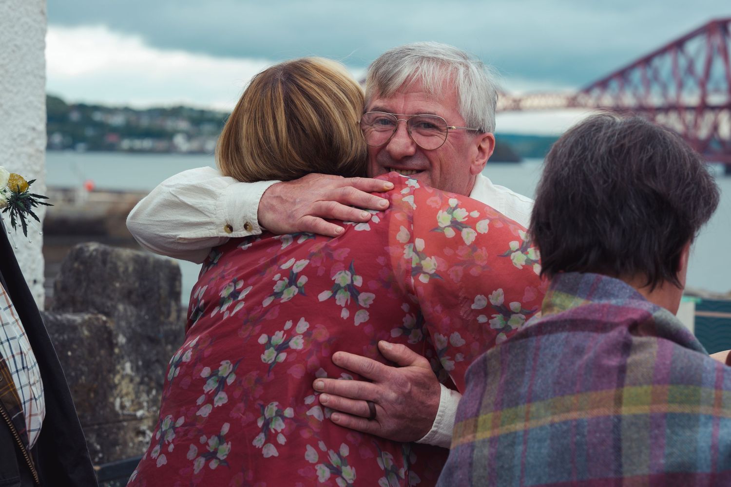Elderly man warmly hugging a woman in a floral dress outdoors near a bridge over water on a cloudy day.