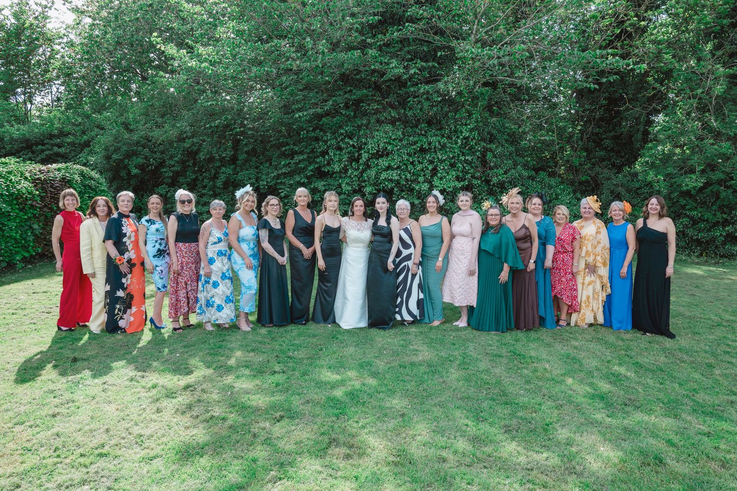 Large group of elegantly dressed women posing together outdoors on green lawn with lush trees in background.