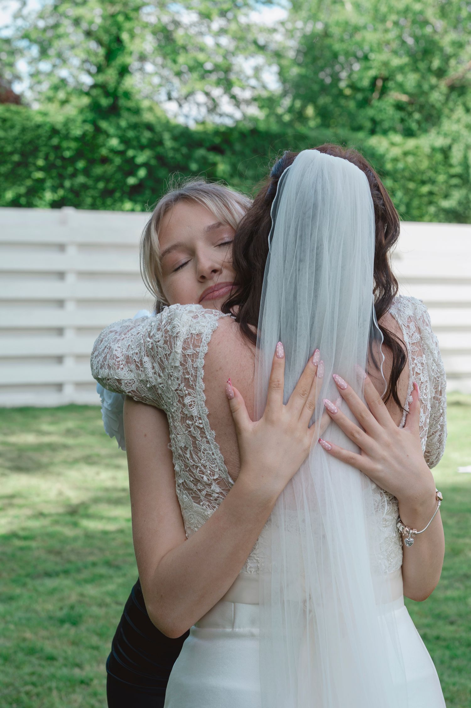 A bride in a white gown and veil shares a heartfelt hug with a blonde woman in a lace dress outdoors.