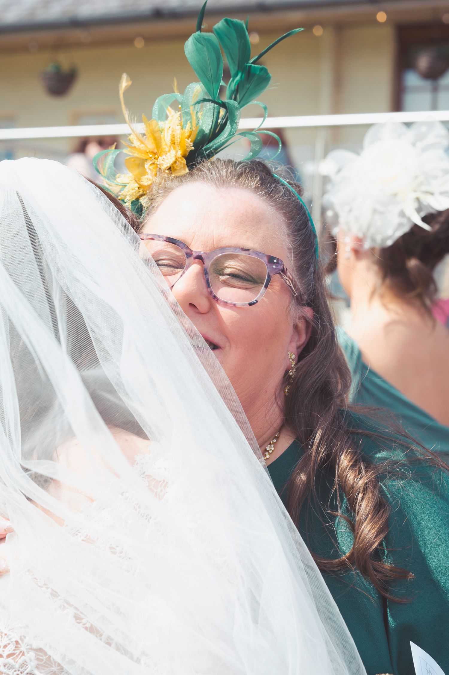 Woman with green fascinator and pink glasses kisses bride through wedding veil at outdoor ceremony.