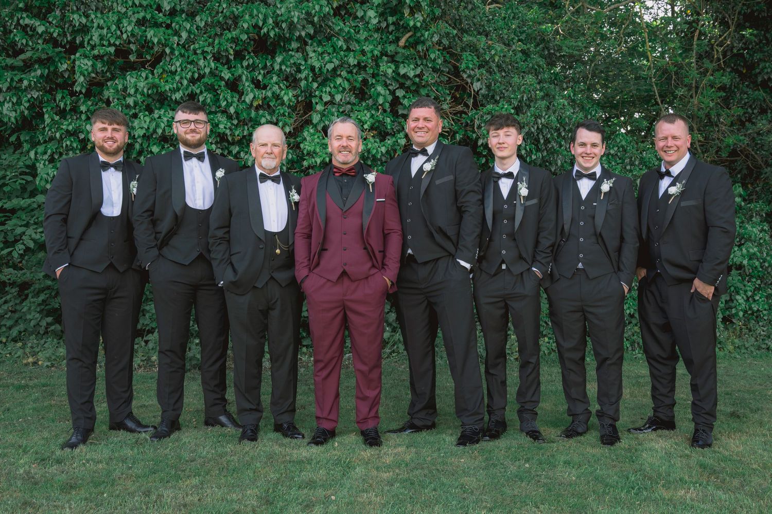 Groom in burgundy suit stands with seven groomsmen in black tuxedos outdoors against lush green ivy backdrop.