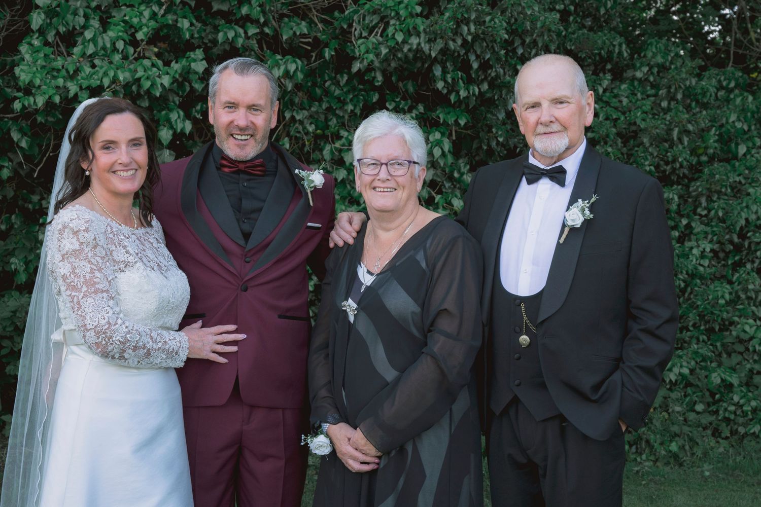 A bride and groom pose with an older couple at a wedding, all dressed formally, outdoors with green foliage background.