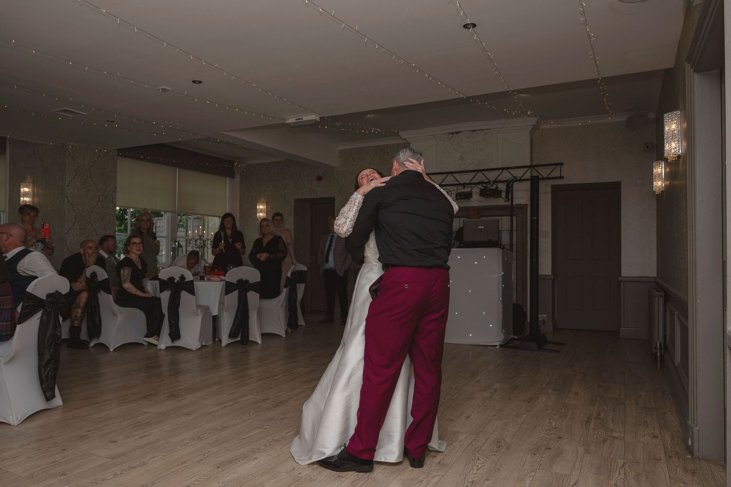 Couple sharing their first dance at a wedding reception, bride in white gown, groom in burgundy pants, guests watching.