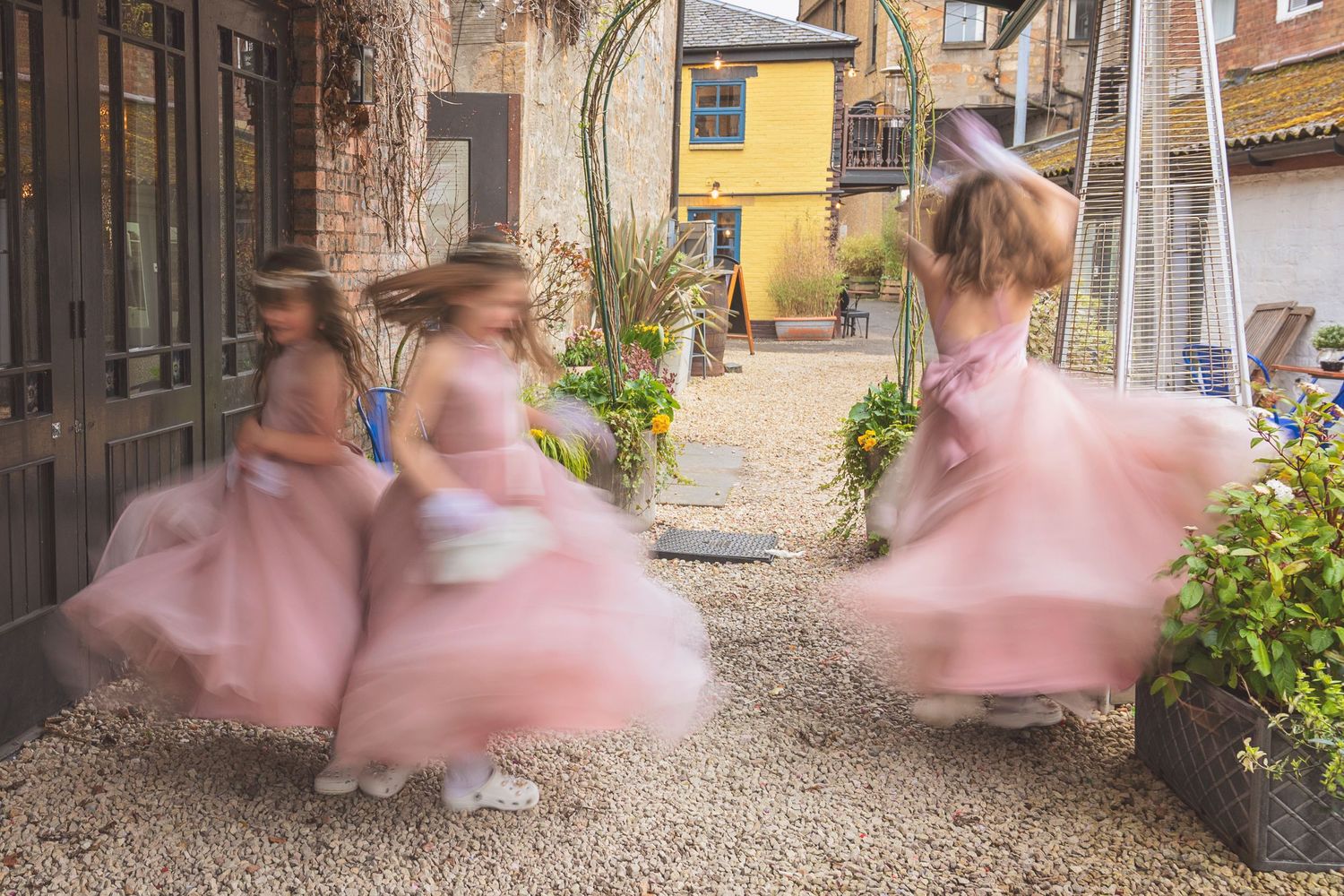 Three flower girls in pink tulle dresses spinning joyfully in a courtyard at a wedding.