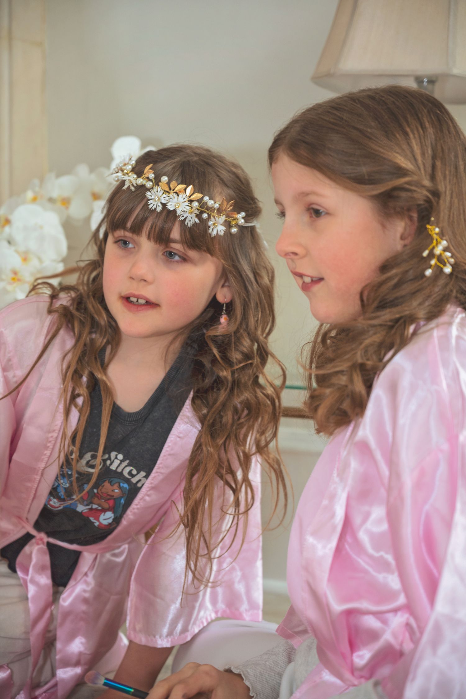 Two young flower girls in pink satin robes wearing floral hair accessories at a wedding preparation.