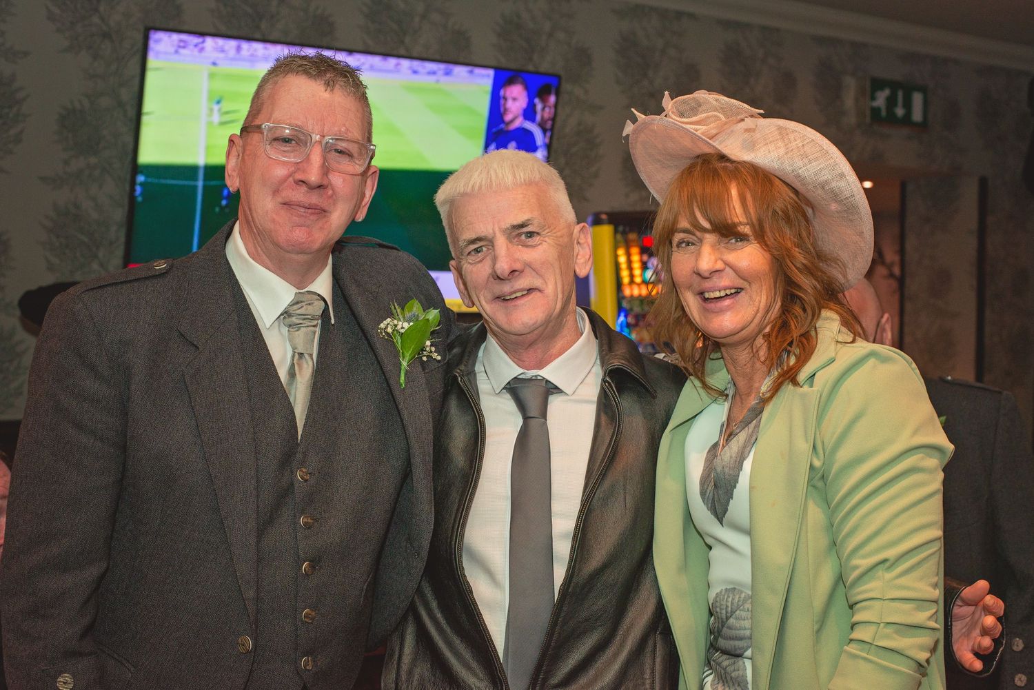 Three guests in formal attire smile at a wedding reception with a TV screen visible in background.