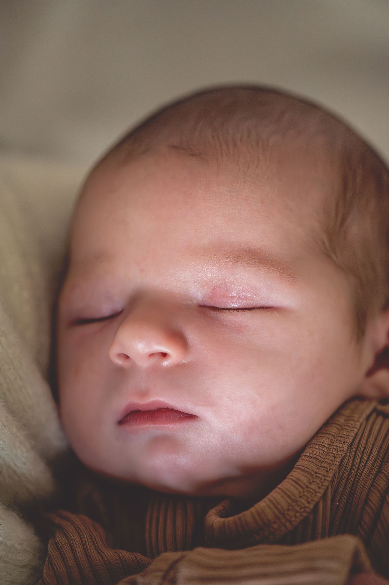 Close-up view of a sleeping baby's face with eyes closed, wrapped in a brown knit blanket.