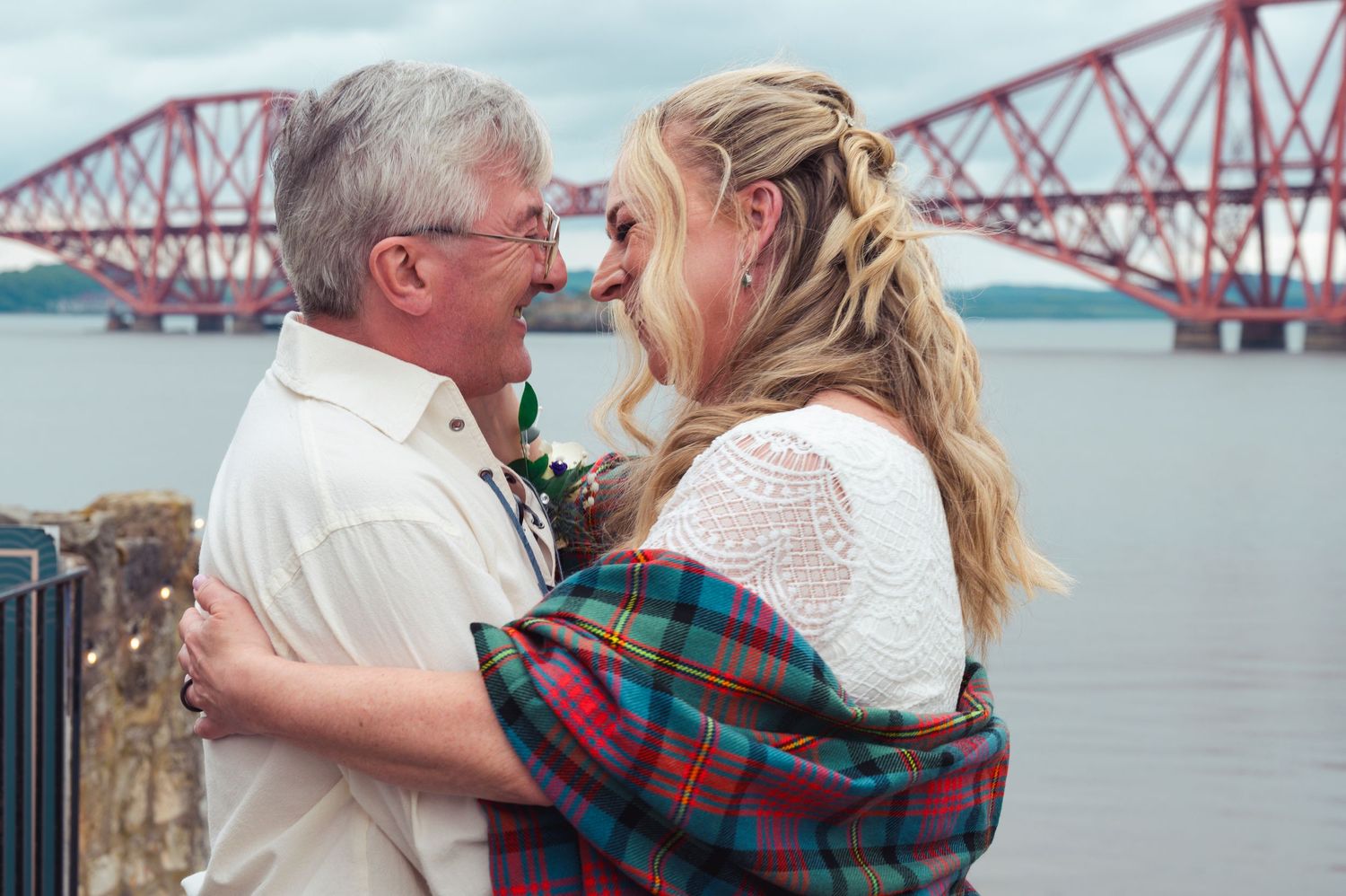 Couple embracing romantically by the Forth Bridge in Scotland, woman wrapped in tartan plaid on a cloudy day.
