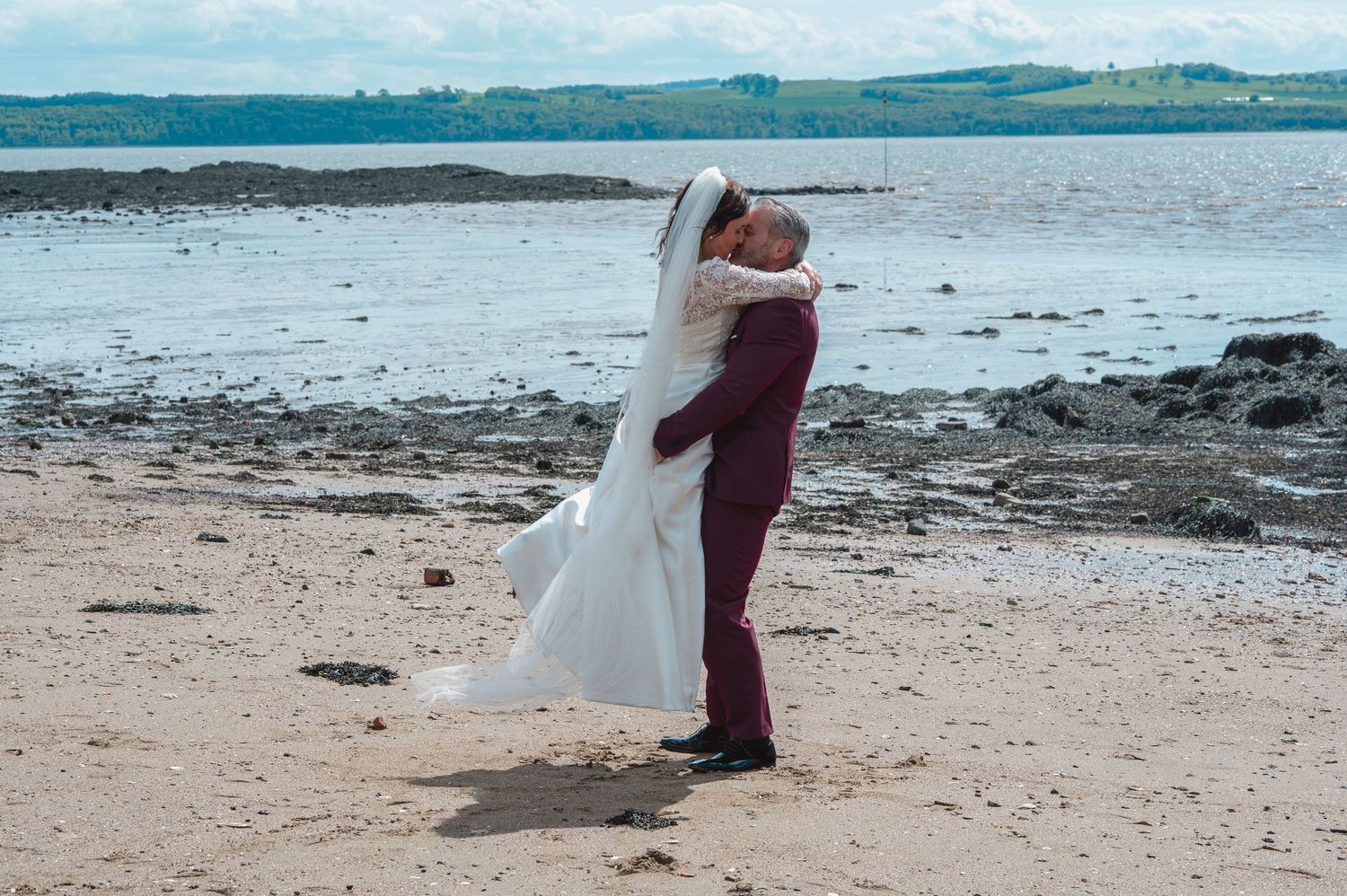A groom in burgundy suit lifts his bride in white wedding dress on a sandy beach by the sea.