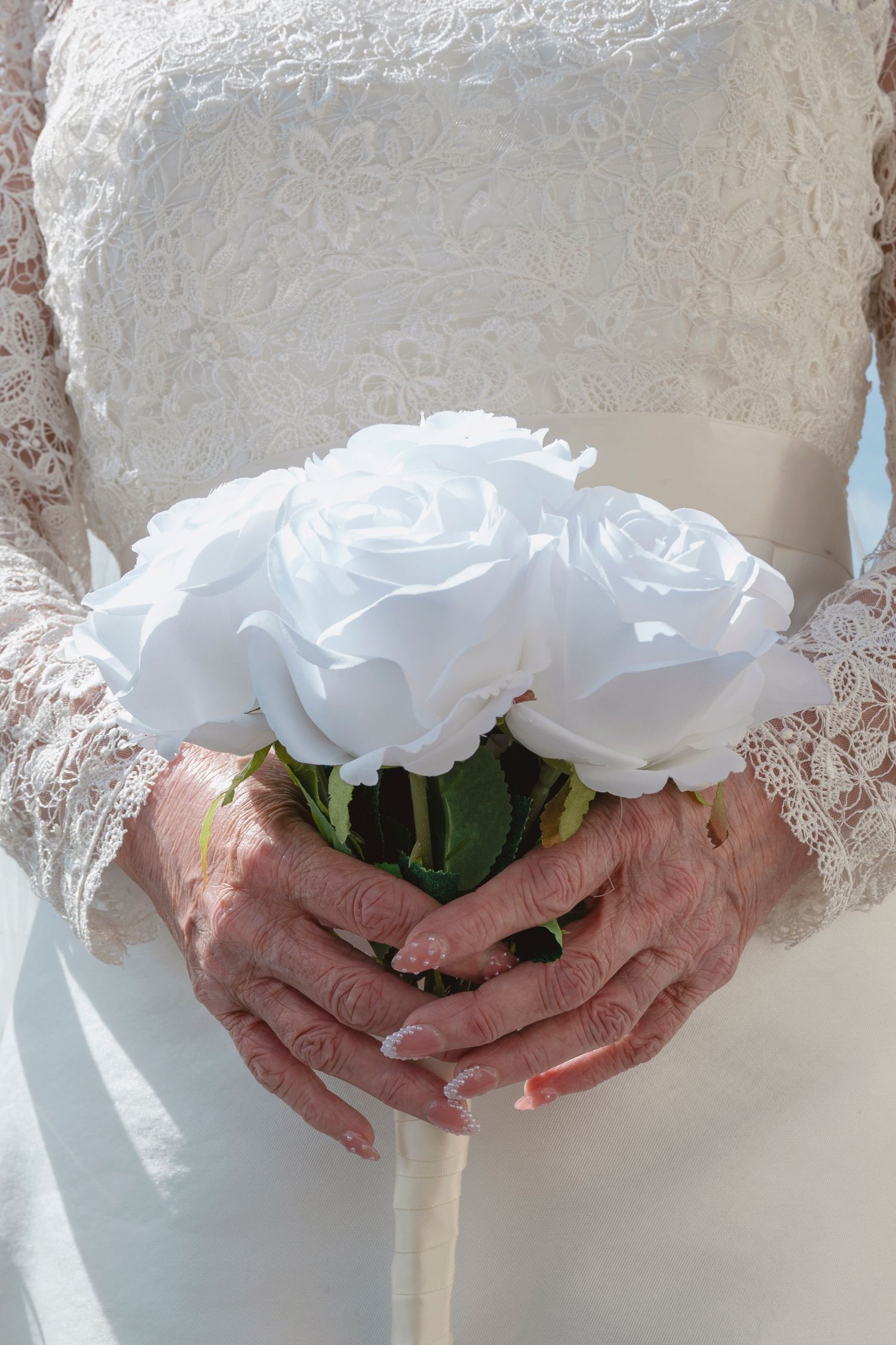 Elderly bride holding white rose bouquet in lace wedding dress, manicured hands visible.