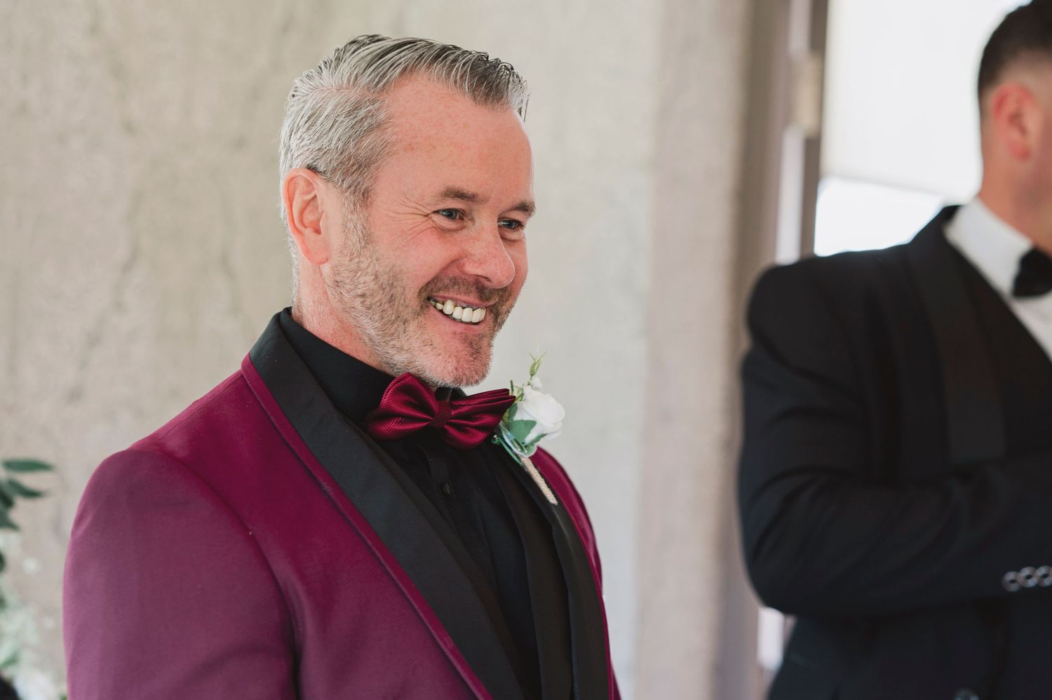 Smiling man in burgundy tuxedo jacket with black bow tie and white boutonniere at a formal event.