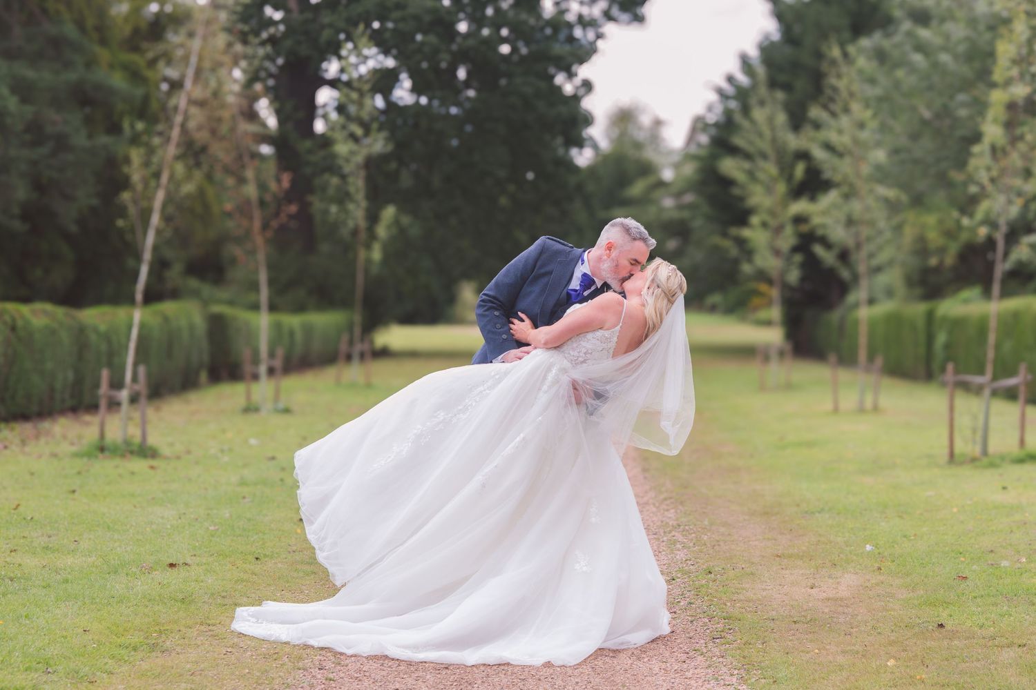 Groom dips bride for a romantic kiss on a tree-lined garden path, bride in flowing white gown with veil.