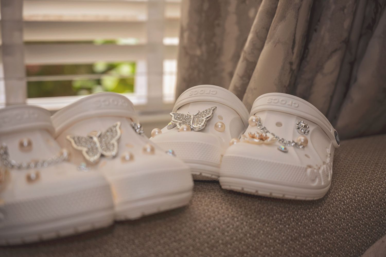 White Crocs decorated with pearl and crystal Jibbitz charms displayed on a chair near a window.