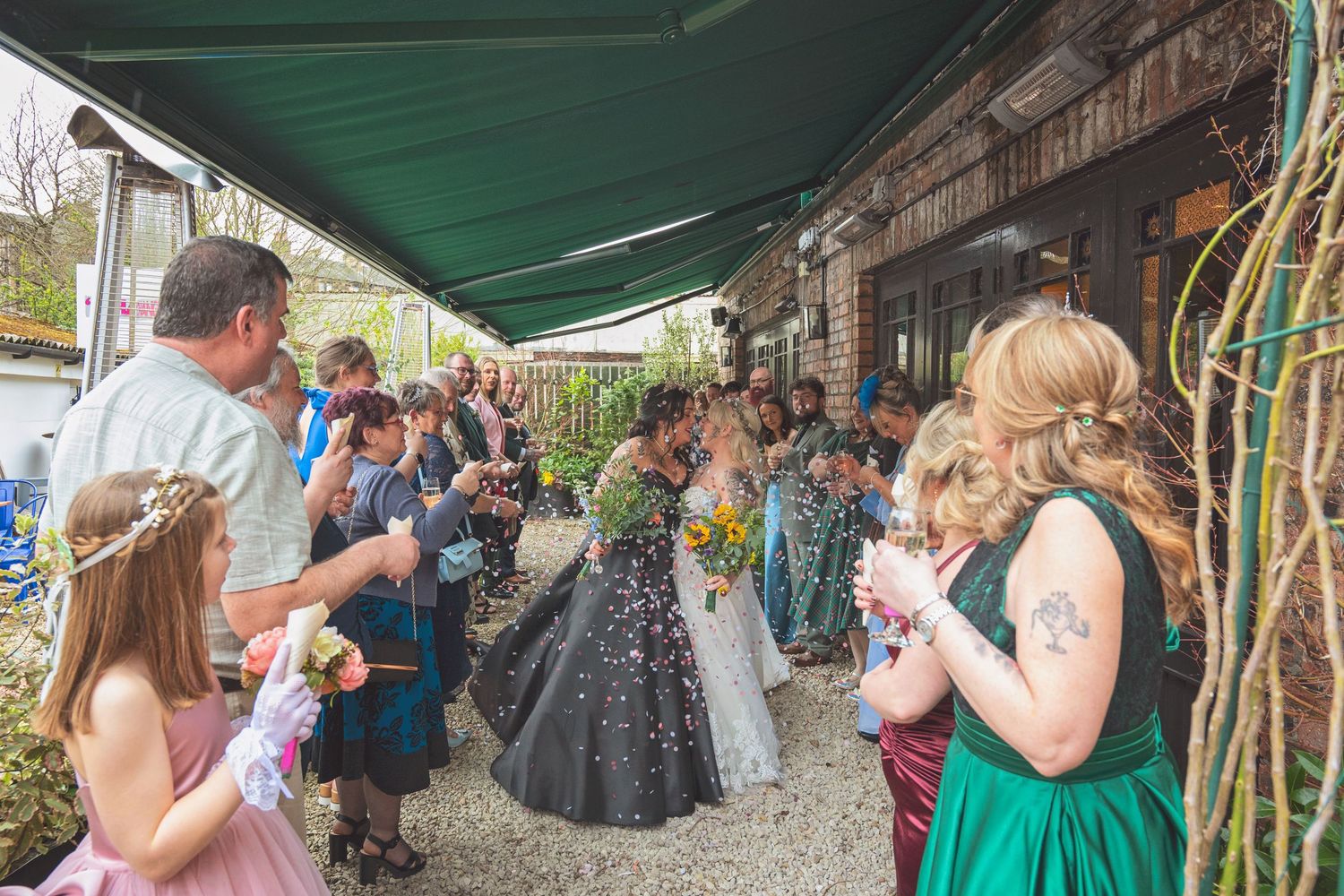 Wedding guests blow confetti as two brides walk through a celebratory tunnel outdoors.