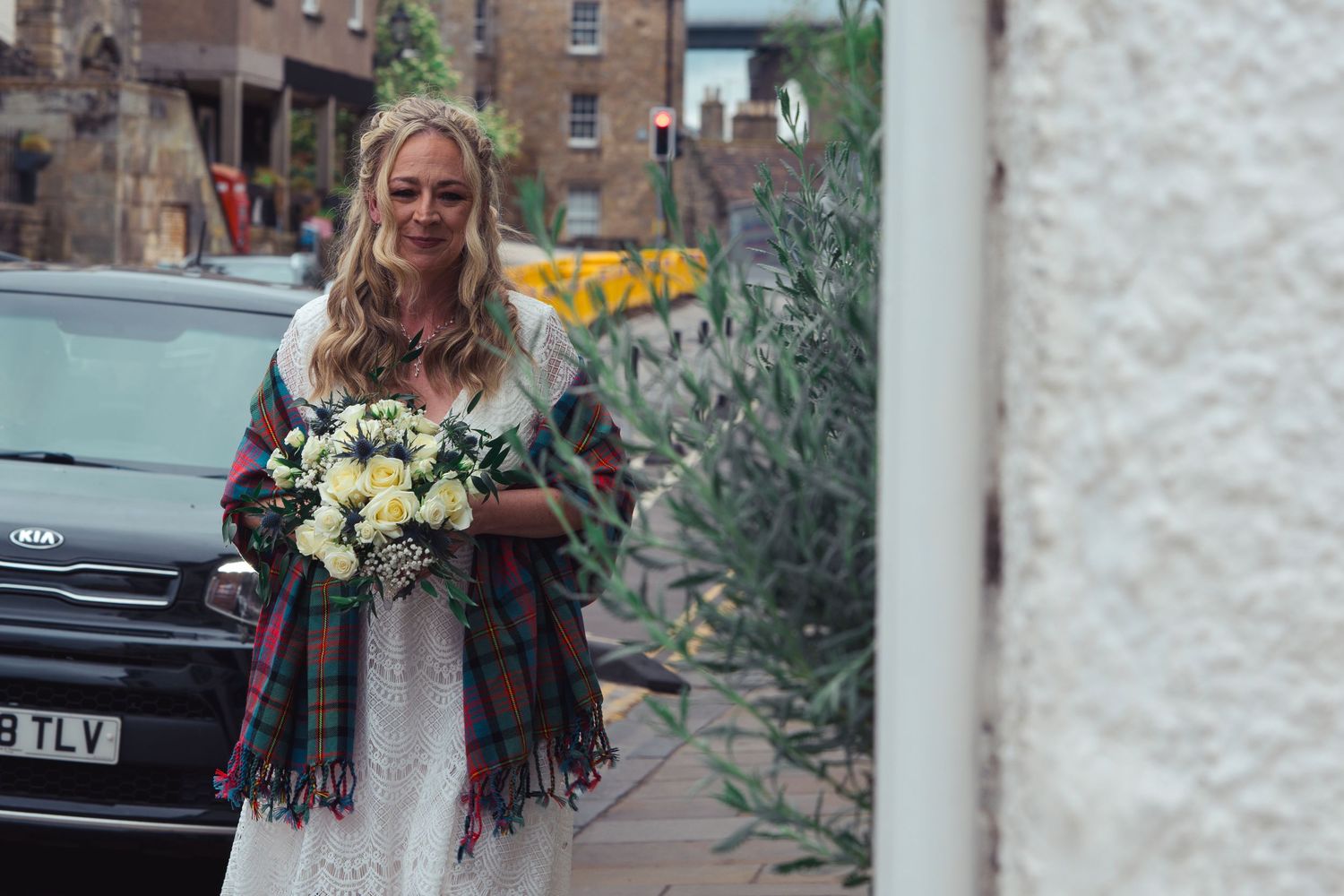 Bride in white lace dress with tartan shawl holding white bouquet outside on wedding day.