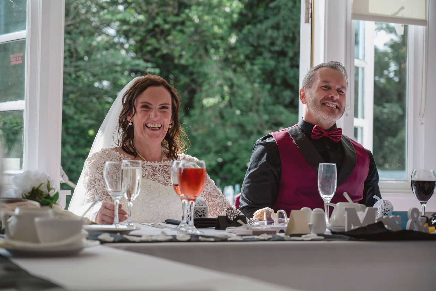Bride and groom laughing joyfully at wedding reception table with wine glasses and elegant white decor.