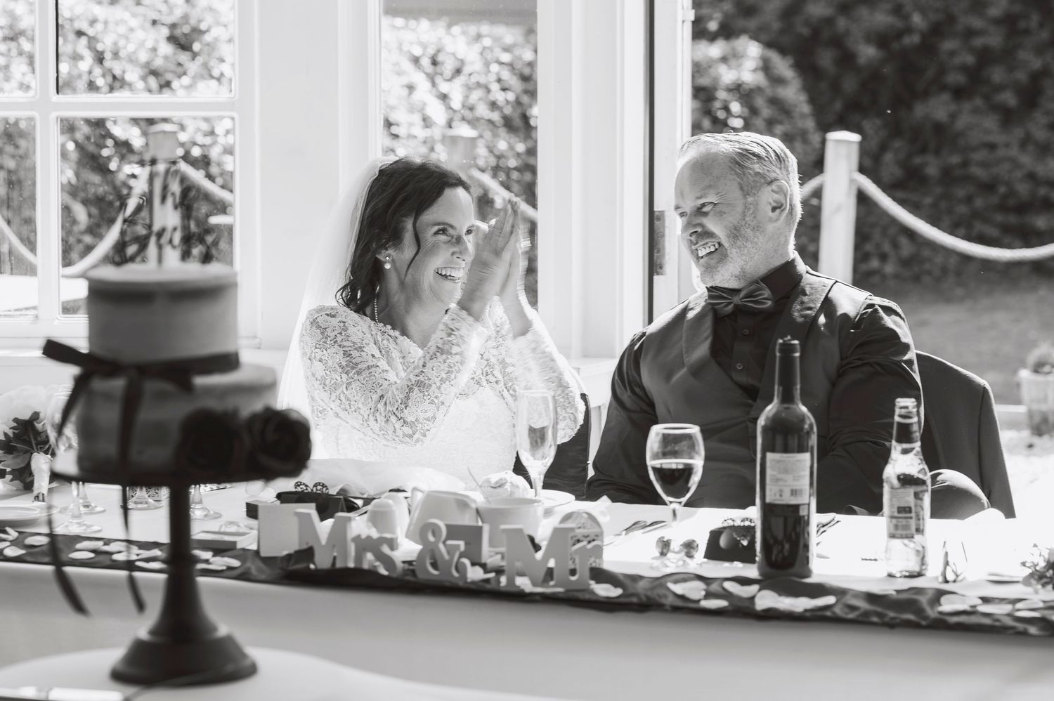 Bride and groom laughing together at wedding reception table with wine bottles and glasses in black and white photo.