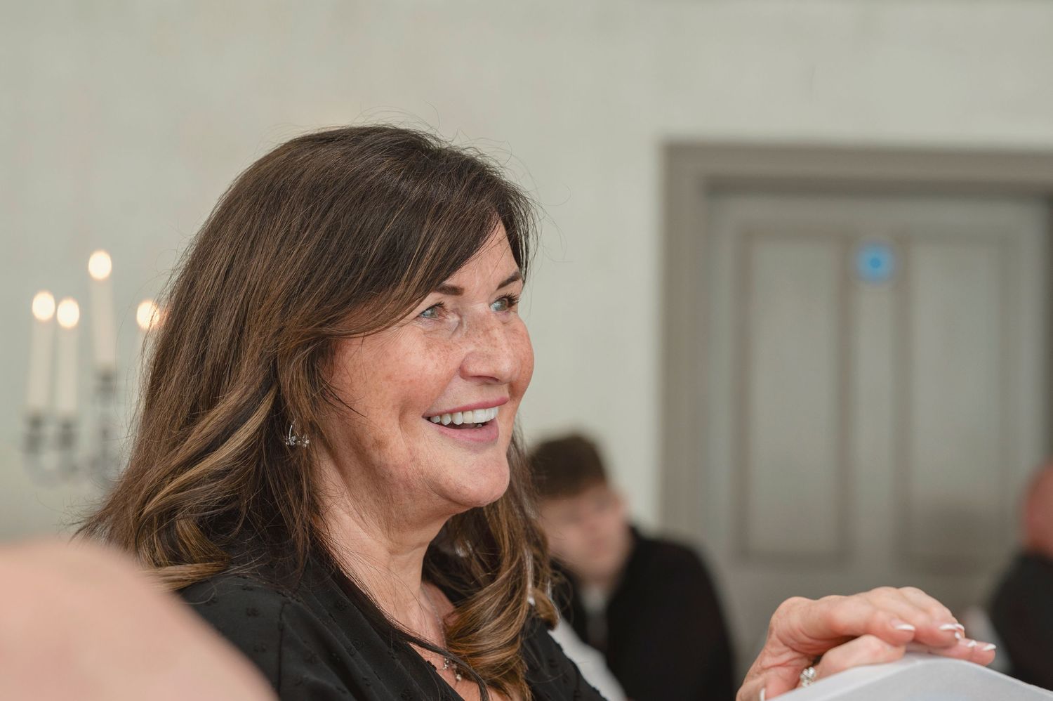 Smiling middle-aged woman with brown hair at an indoor event, wearing a dark jacket.