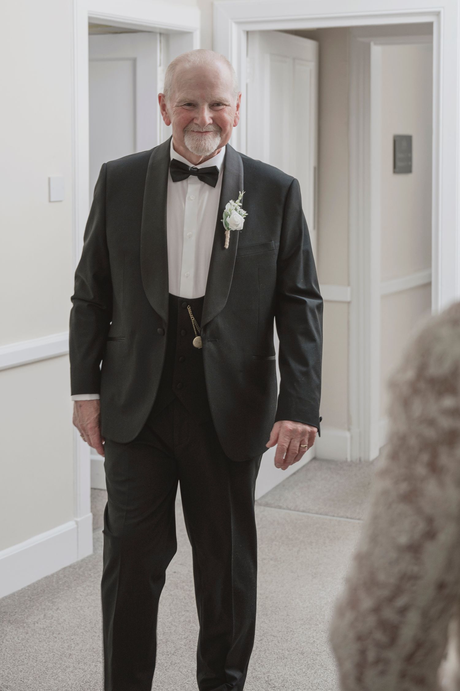 Elderly man in black tuxedo with bow tie and white boutonniere smiling in hallway at formal event.
