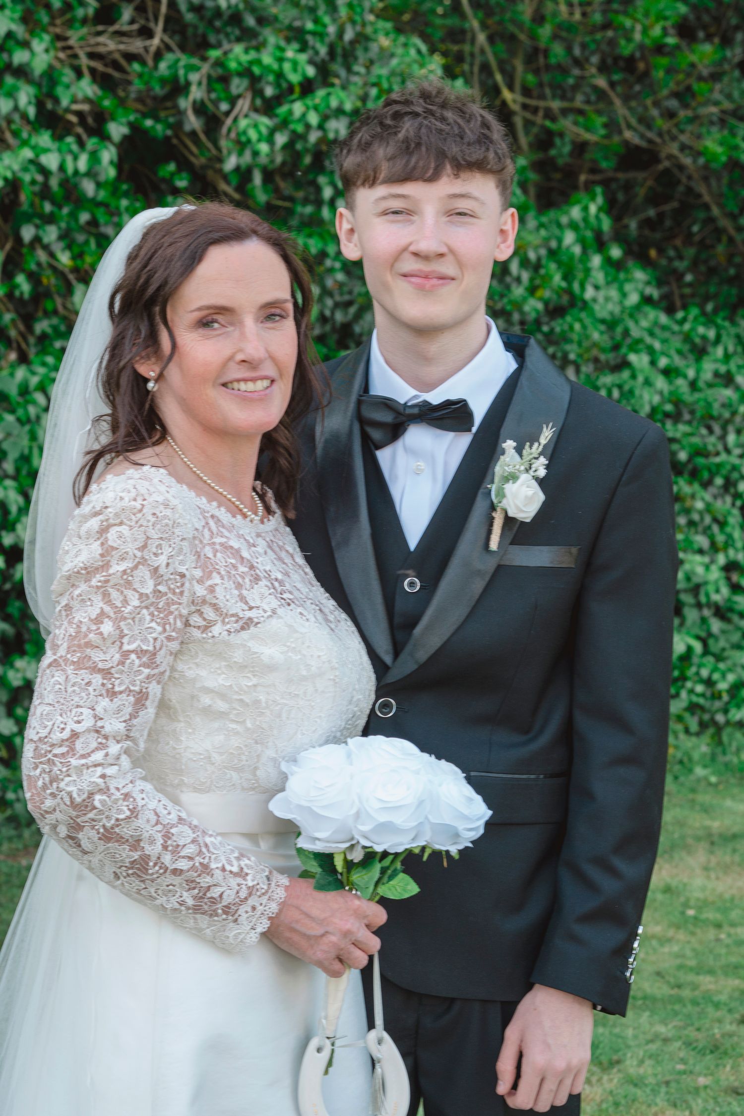 Bride in lace gown holding white bouquet poses with young man in black tuxedo at outdoor wedding ceremony.