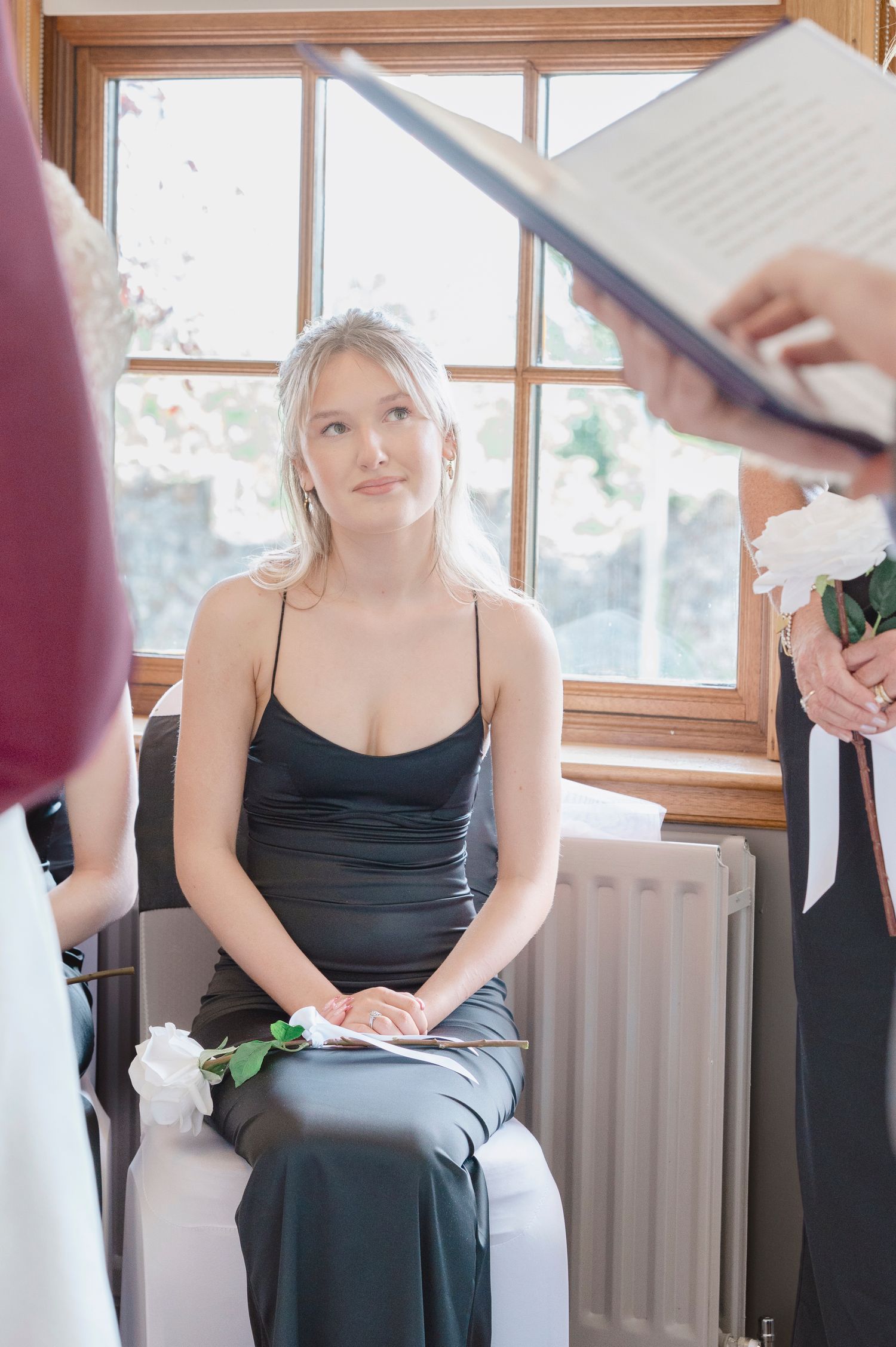Young woman in black satin dress seated at wedding ceremony, looking up attentively near window.