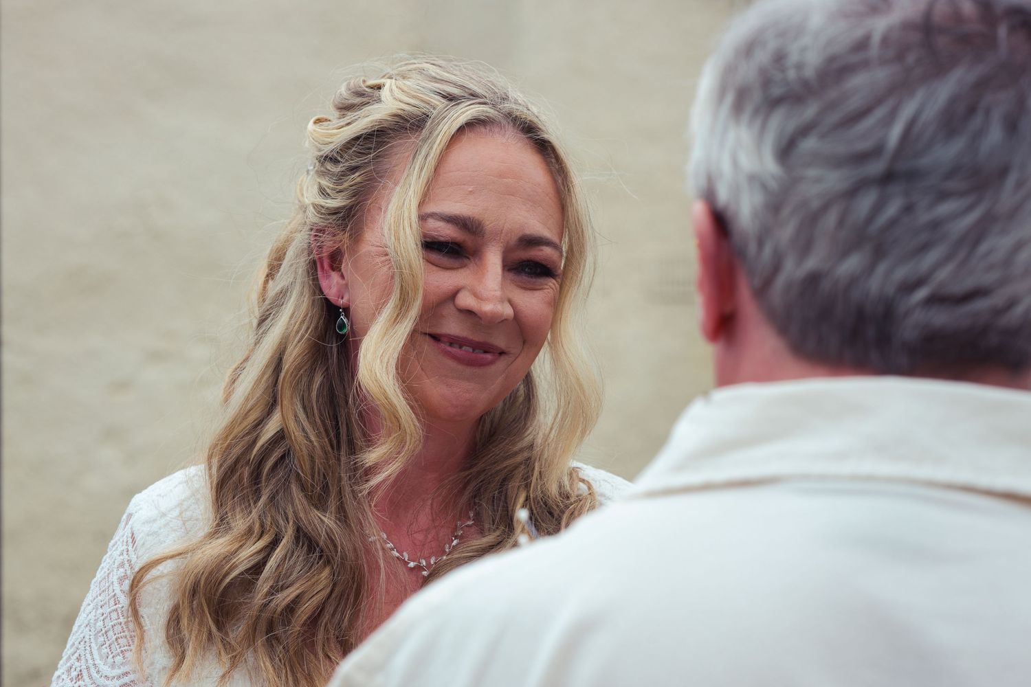Bride smiling warmly at groom during outdoor wedding ceremony, wearing white lace dress and pearl necklace.
