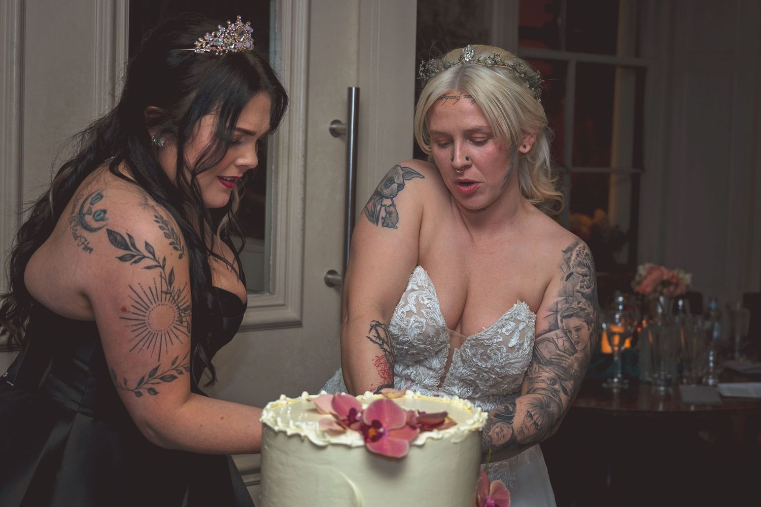 Two tattooed women, one in a tiara and black dress, cut a white wedding cake together.