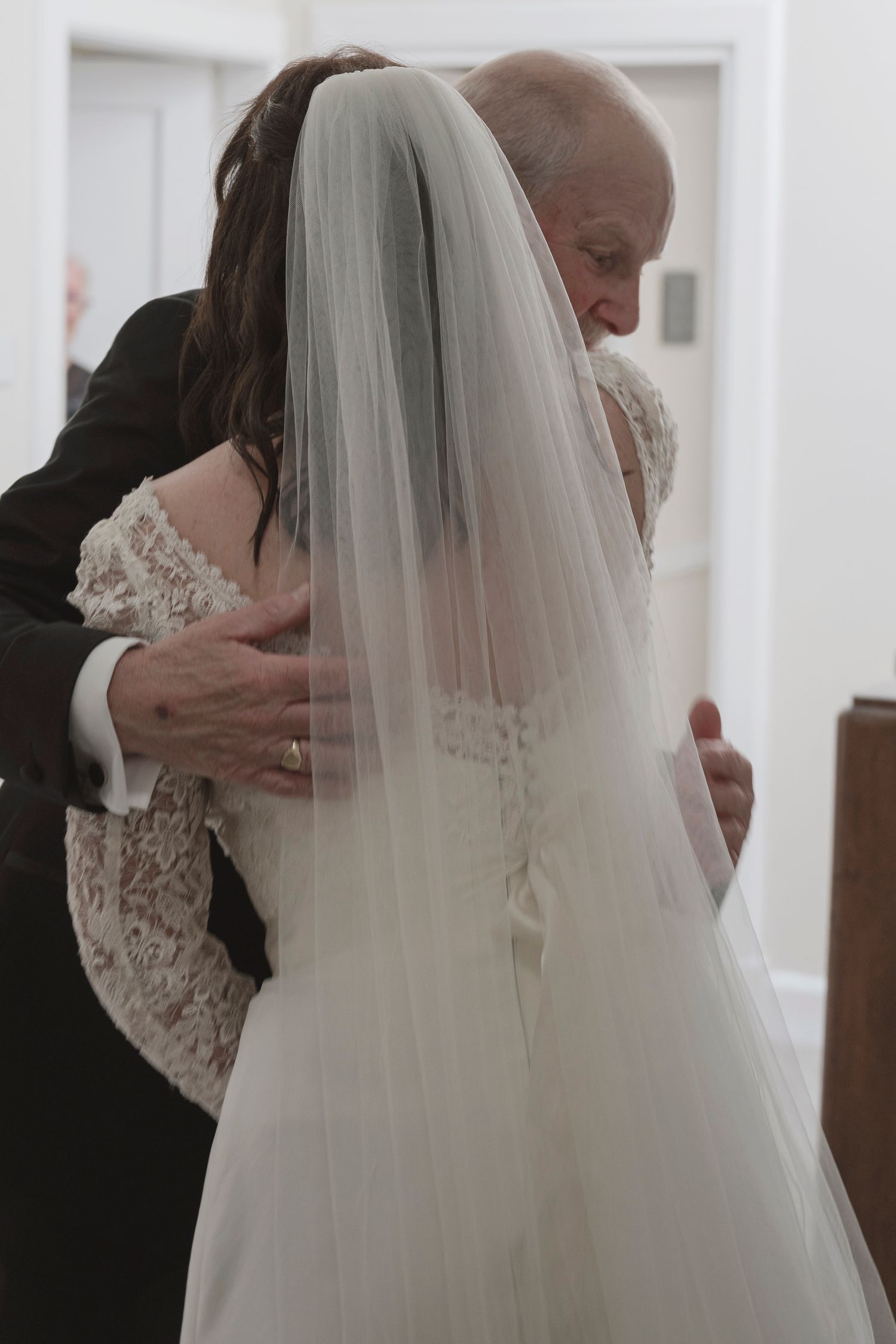 Elderly man embracing bride in white lace wedding dress and veil during emotional father-daughter moment.