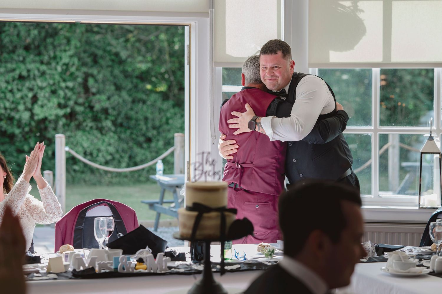 Two men sharing a warm embrace hug at a wedding reception dinner in a bright venue.