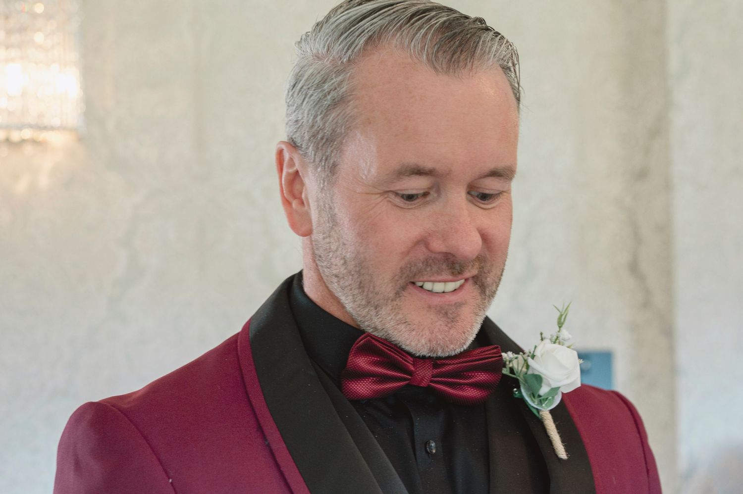 Man in burgundy tuxedo jacket with black bow tie and white boutonniere, smiling at a formal event.