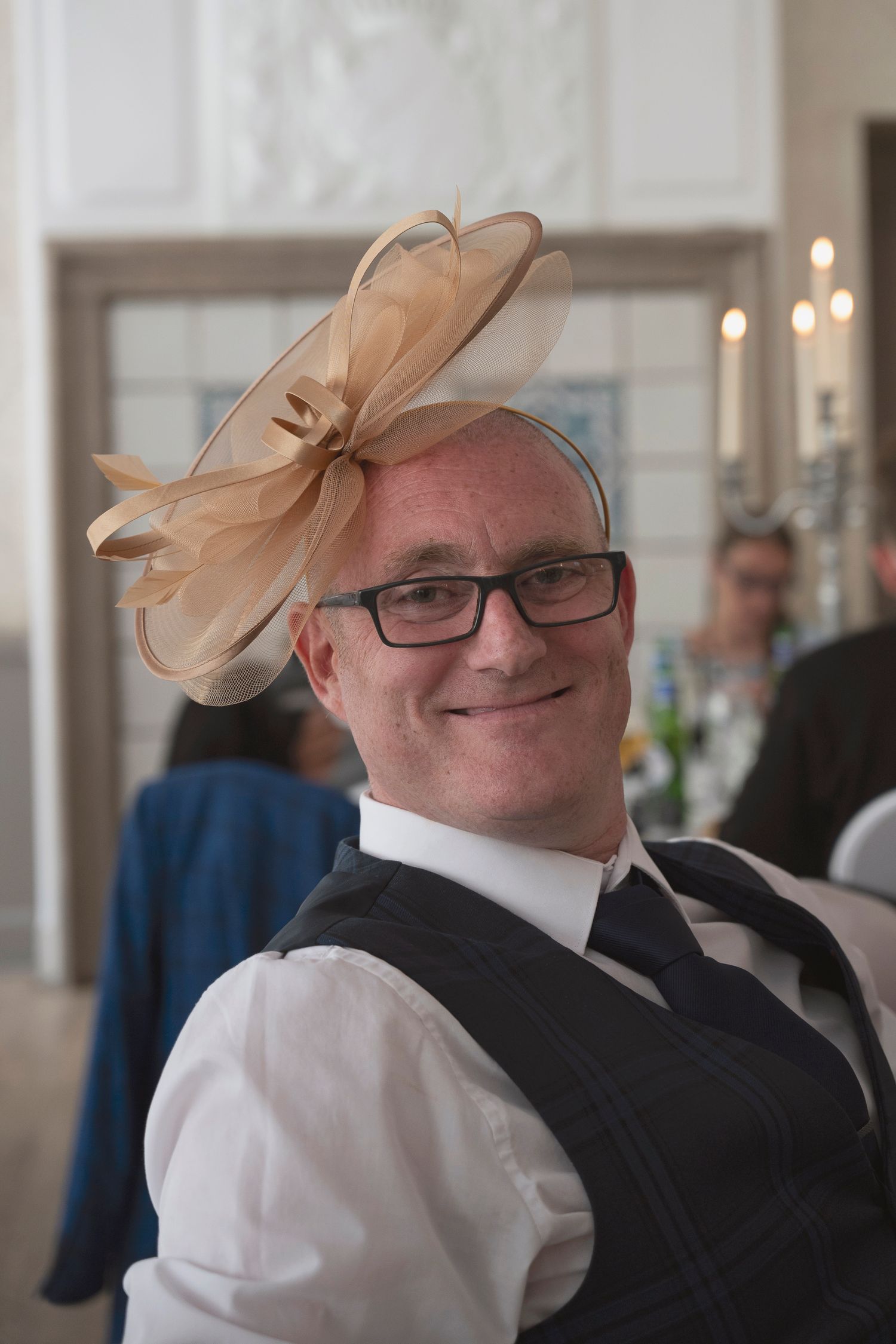 A smiling man wearing glasses and a gold fascinator hat at a formal event.