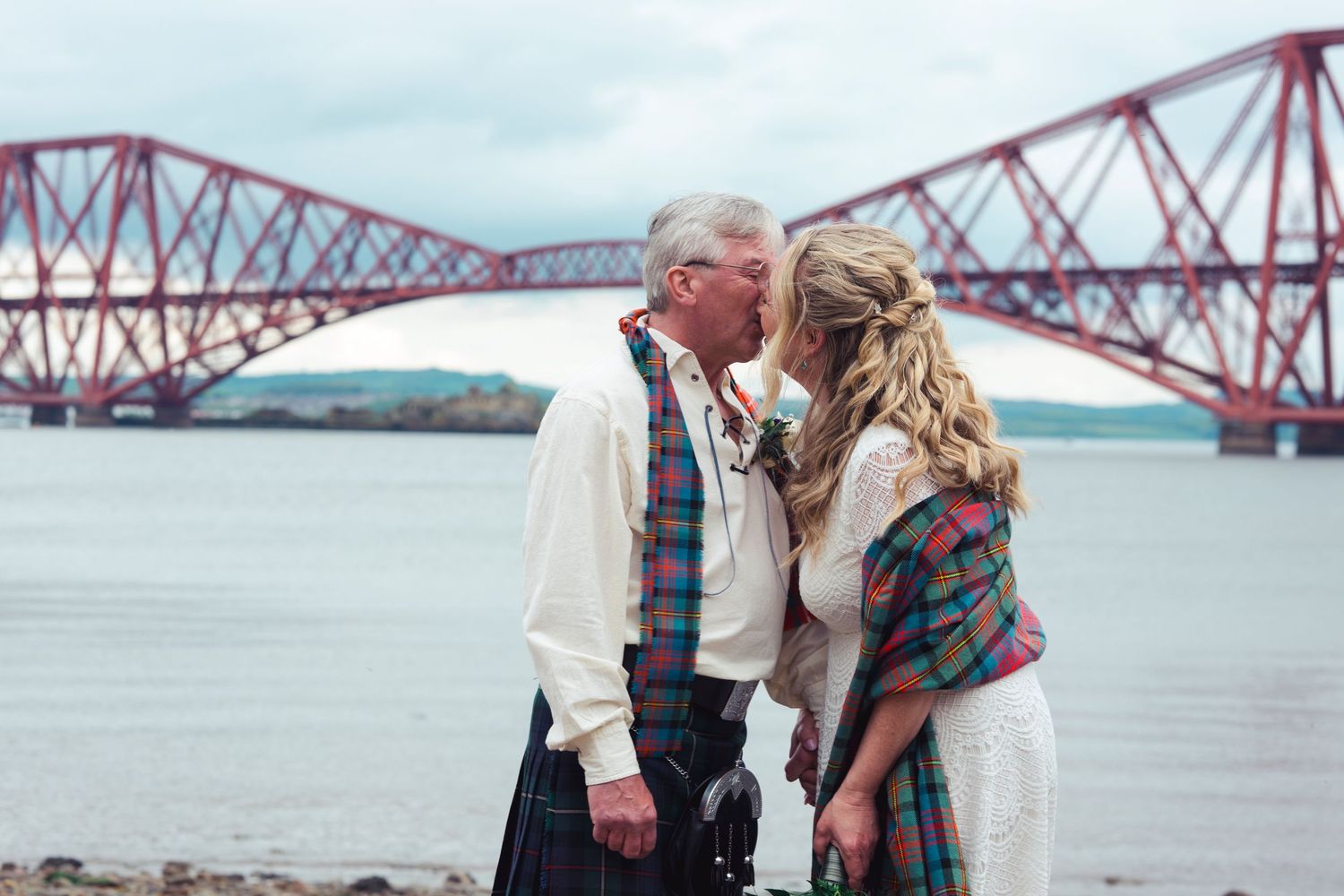 Couple in Scottish attire kissing near the Forth Bridge, Edinburgh, with tartan and kilt in a romantic outdoor setting.