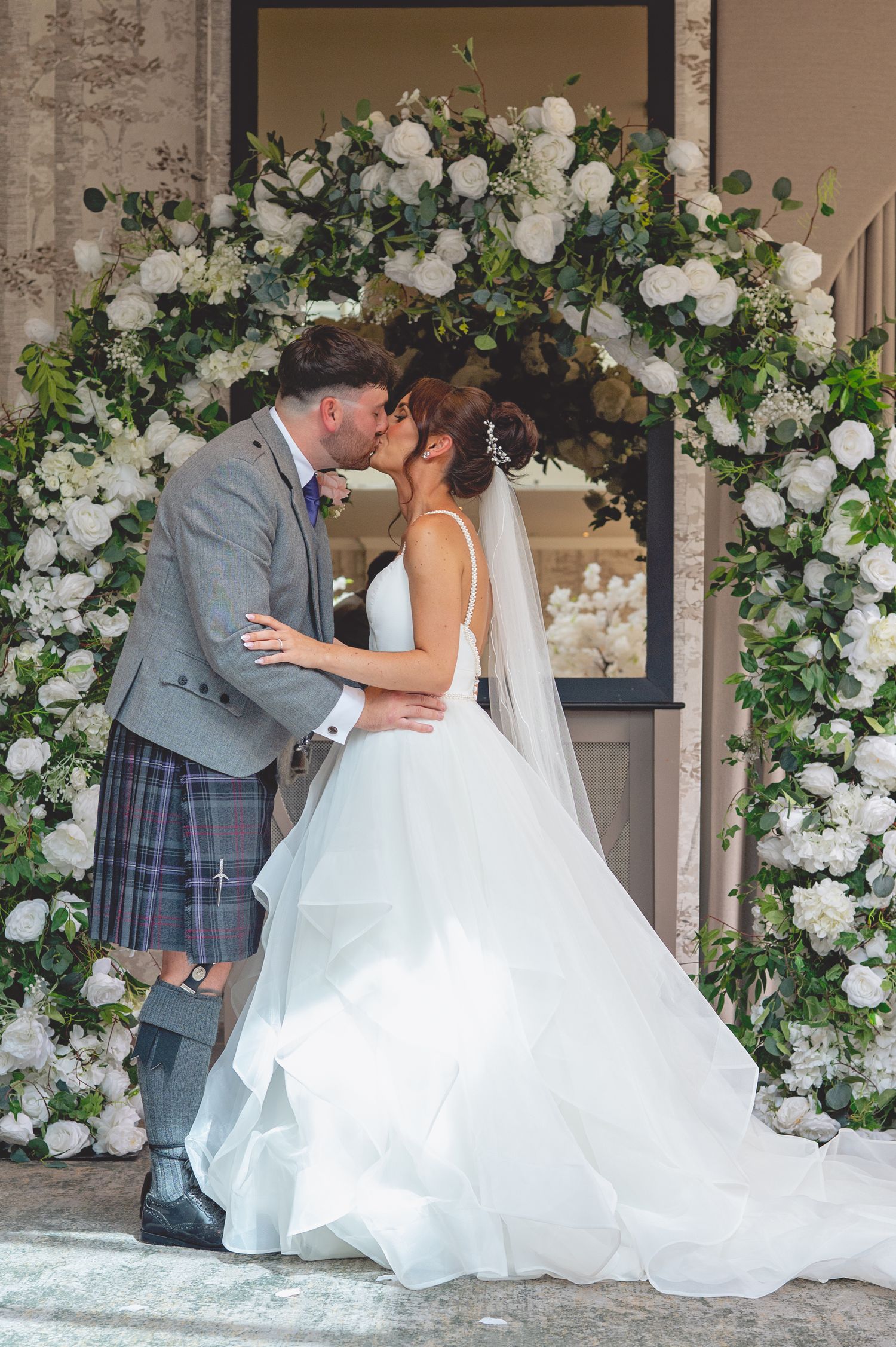 Romantic wedding kiss under an elegant white floral arch at a stone venue.