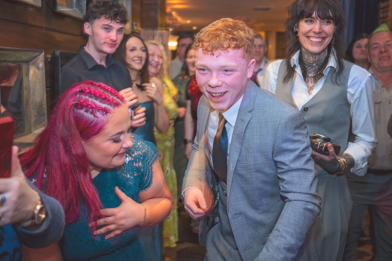 A red-haired teenage boy dances joyfully with guests at a lively wedding reception.