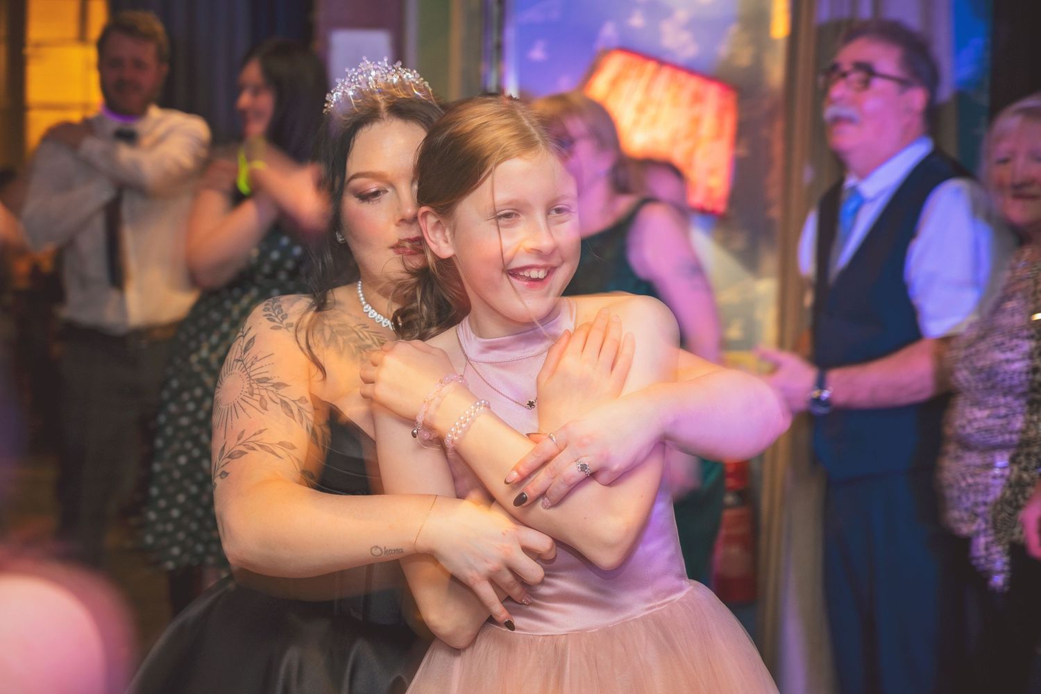 Tattooed bride in black dress dancing joyfully with young girl in pink at wedding reception.